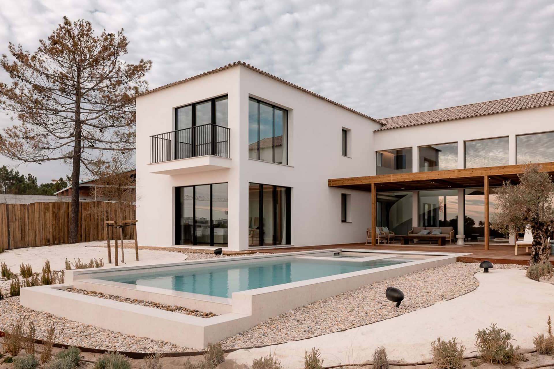 Modern white villa exterior with rectangular swimming pool, wooden pergola, outdoor lounge area, and gravel landscaping under a cloudy sky.