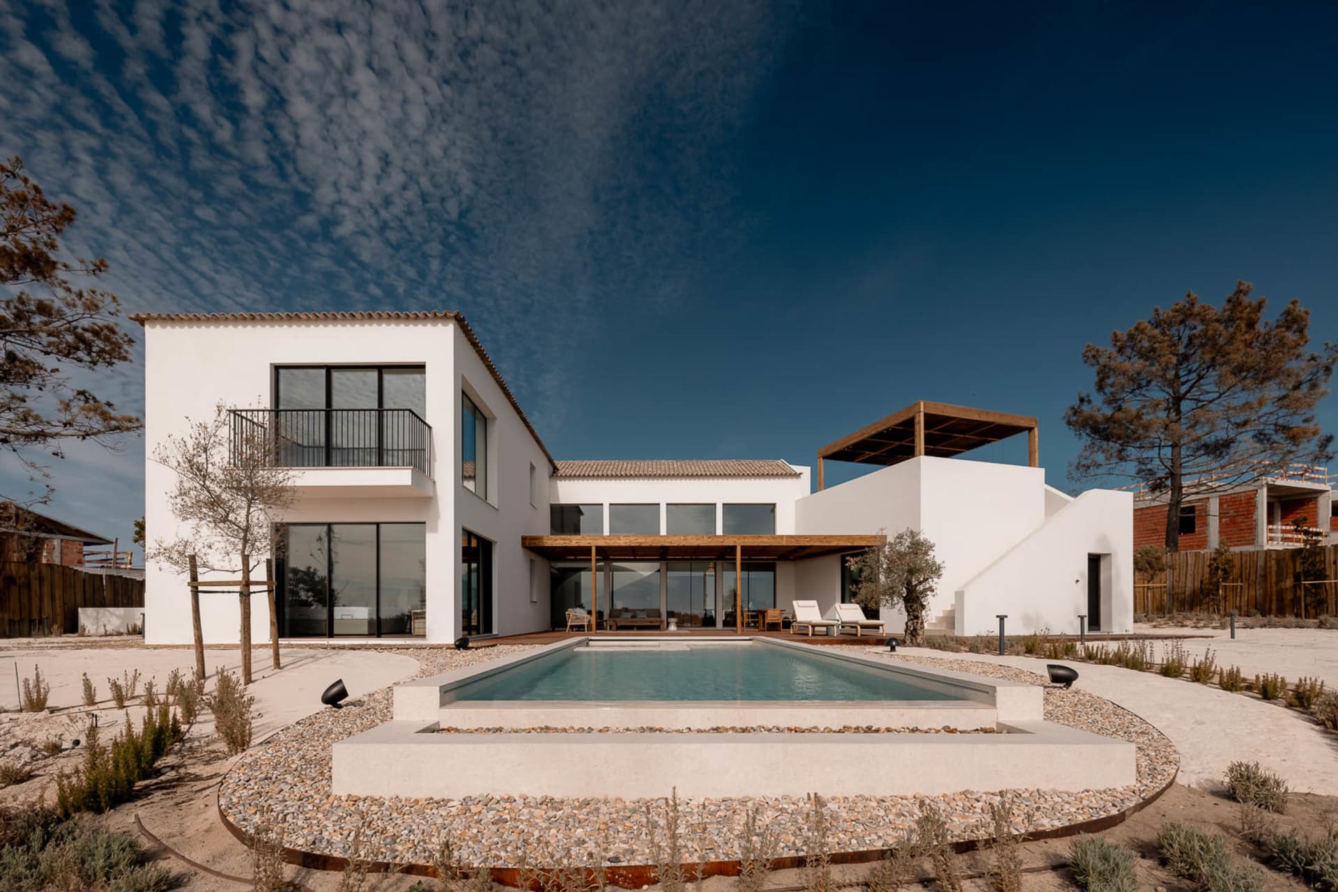 Modern white stucco villa with large glass windows, wooden pergola, rectangular swimming pool, and xeriscaping in a Mediterranean landscape under a deep blue sky.