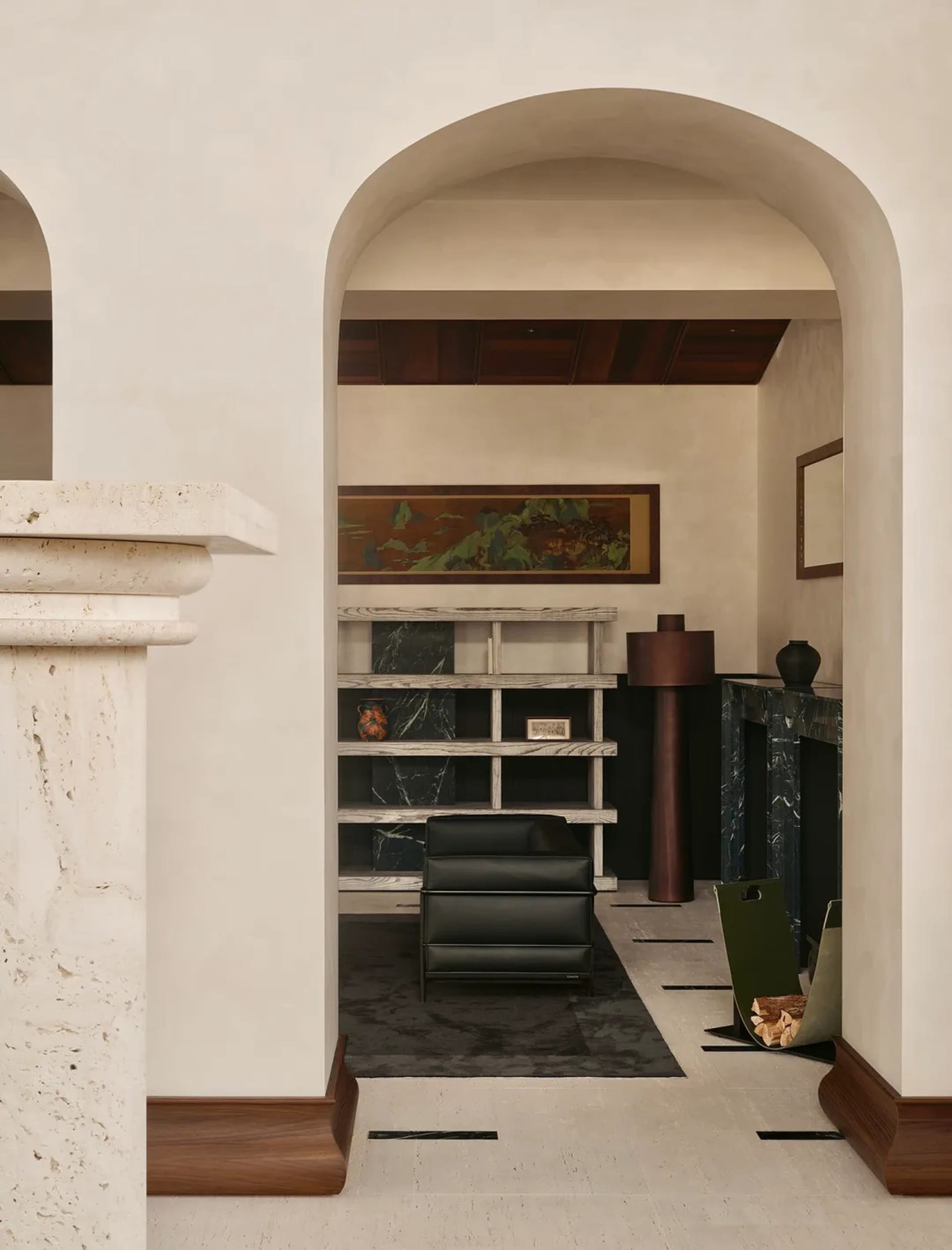 View through a thick beige marble archway into a modern living room featuring a black leather armchair, marble shelving, dark green marble fireplace, and a large landscape painting.