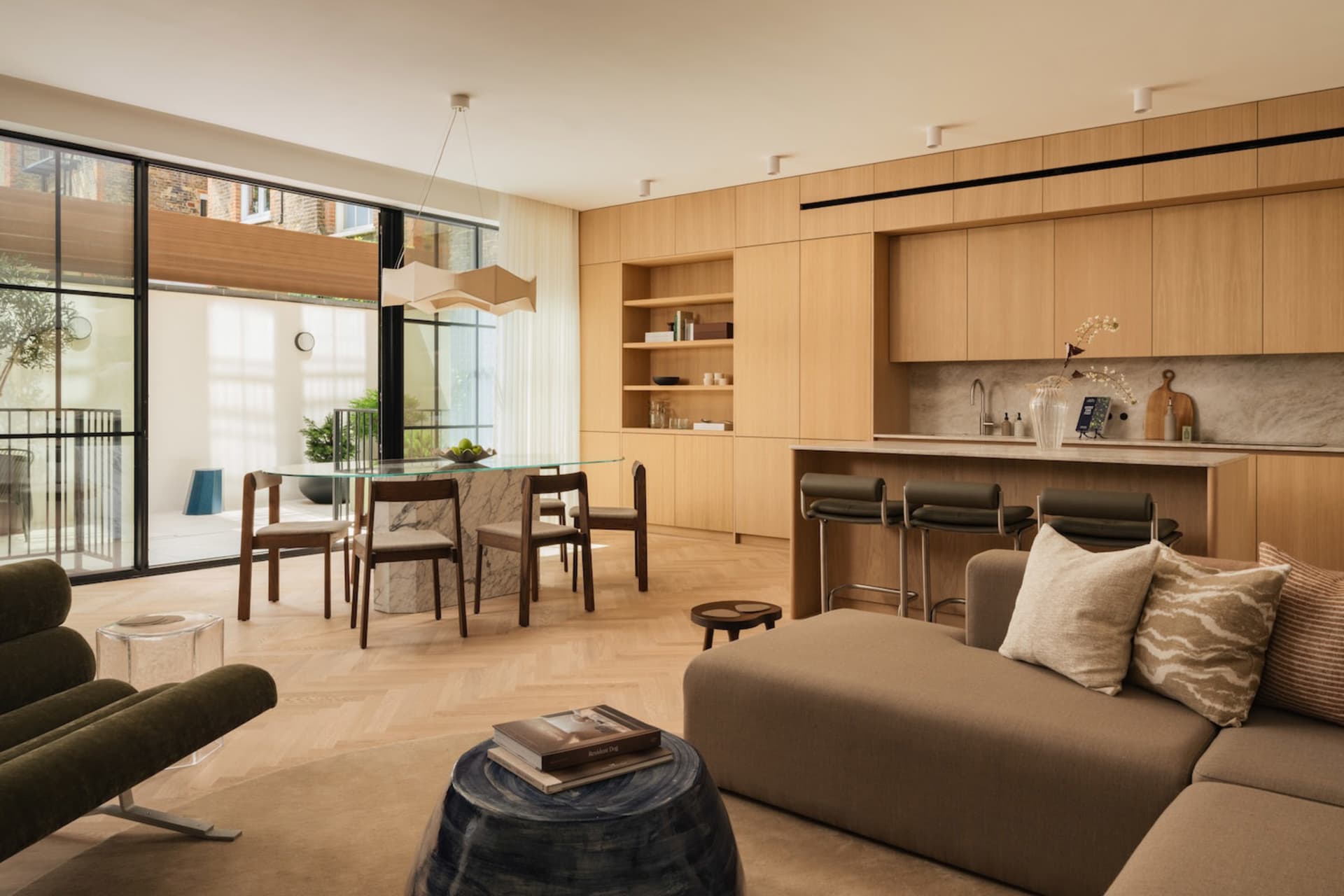 Modern open-plan living room featuring light oak cabinetry, white marble dining table, dark green velvet lounge chair, and a large L-shaped sectional sofa in neutral tones.