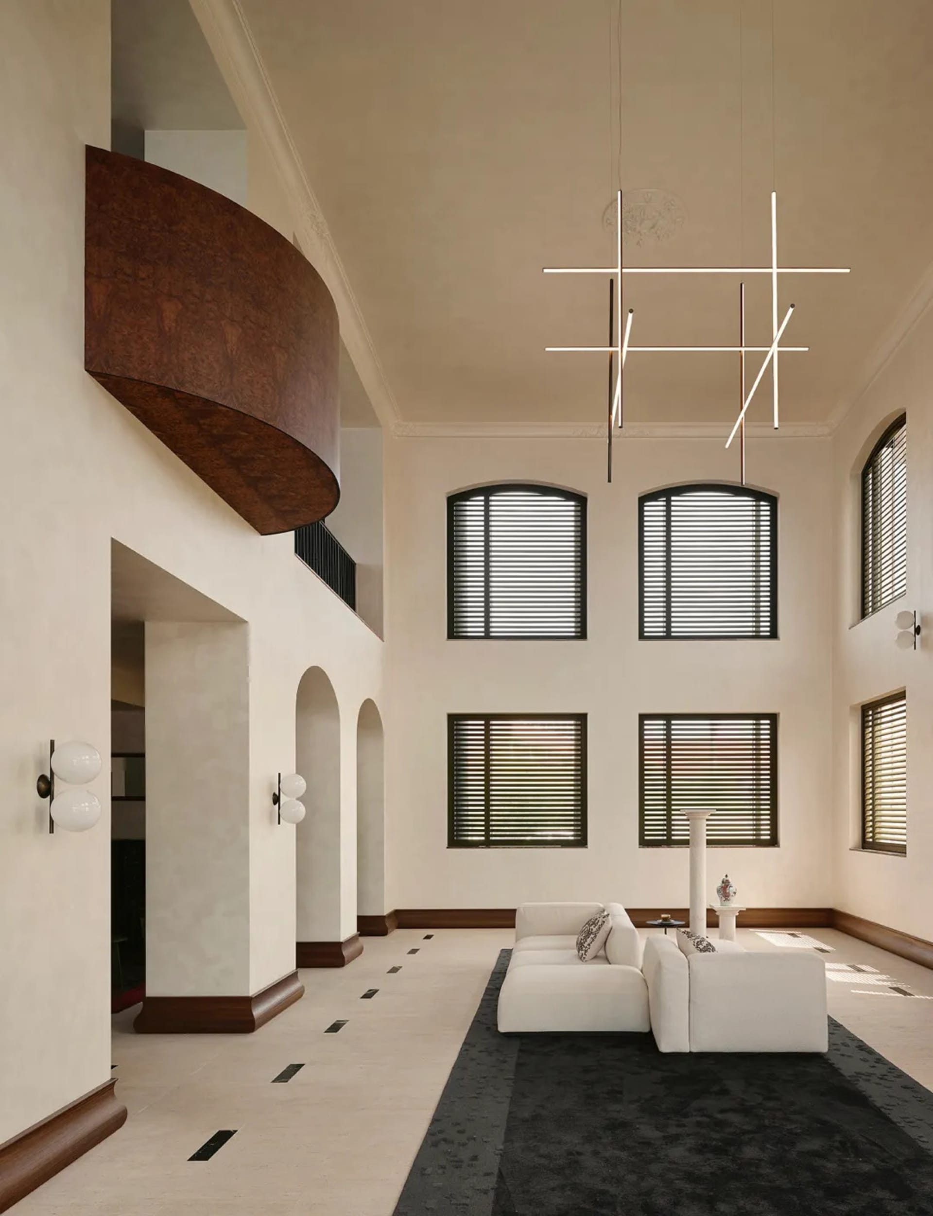 Spacious double-height living room featuring a curved walnut balcony, white L-shaped sectional sofa on a dark rug, modern geometric chandelier, and arched doorways with dark wood trim.