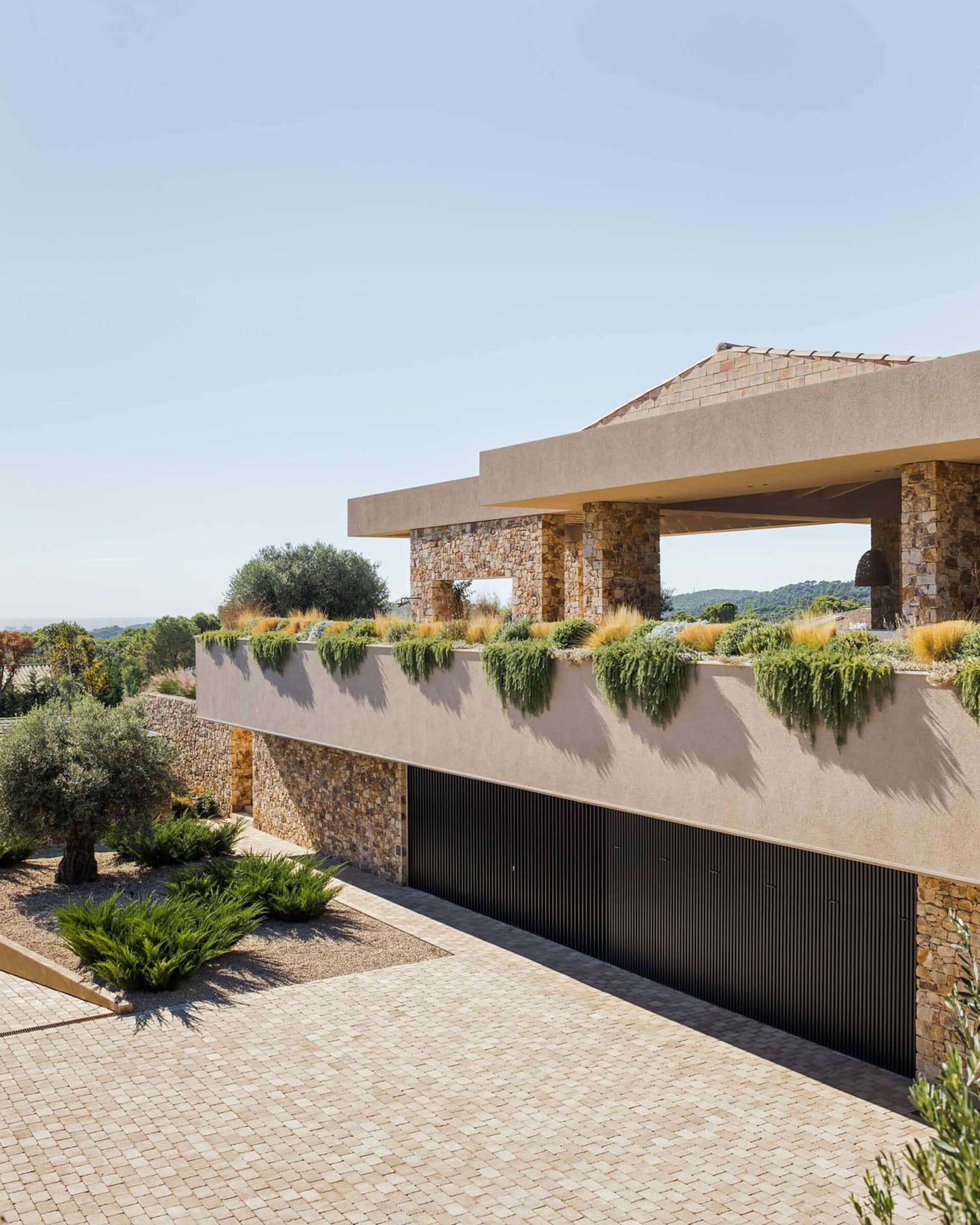 Modern Mediterranean villa exterior with stone walls, concrete overhang, cascading rooftop garden, and paved courtyard under bright sunlight.