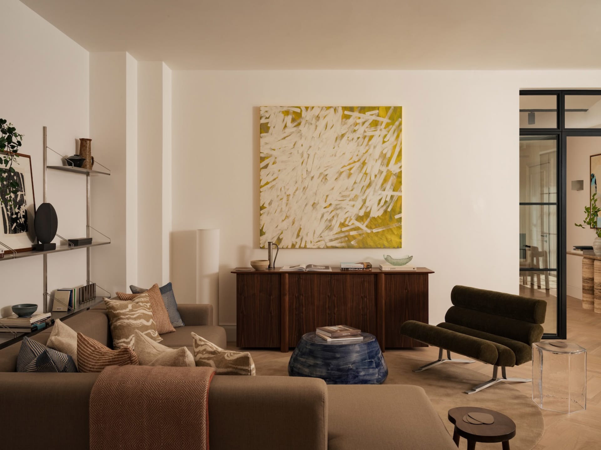 Modern living room featuring a brown sectional sofa with patterned pillows, a dark walnut console table, a large chartreuse and white abstract painting, and a dark green velvet chaise lounge.