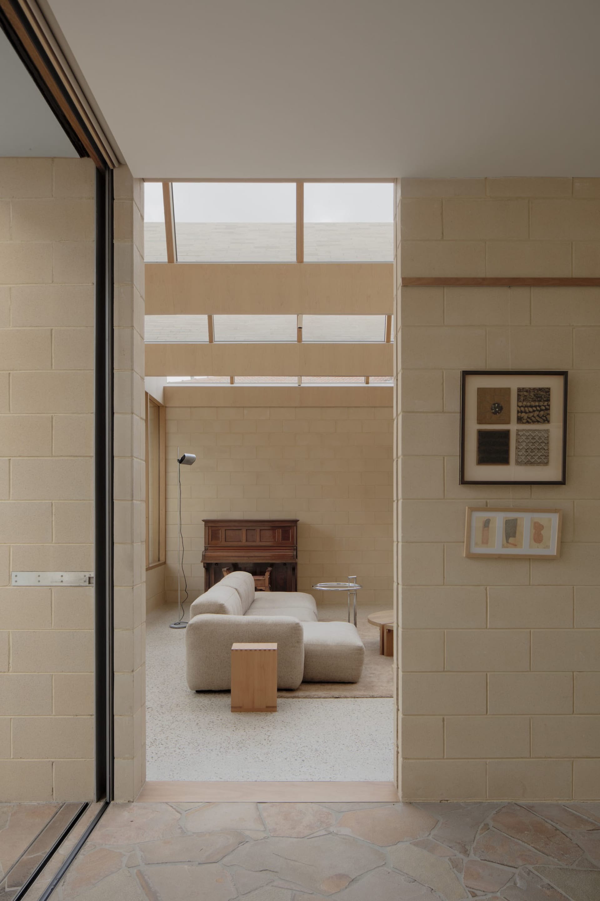 View into a minimalist living room featuring light-colored concrete block walls, a large modular beige sofa on terrazzo flooring, wooden accents, and high clerestory windows allowing soft natural light.