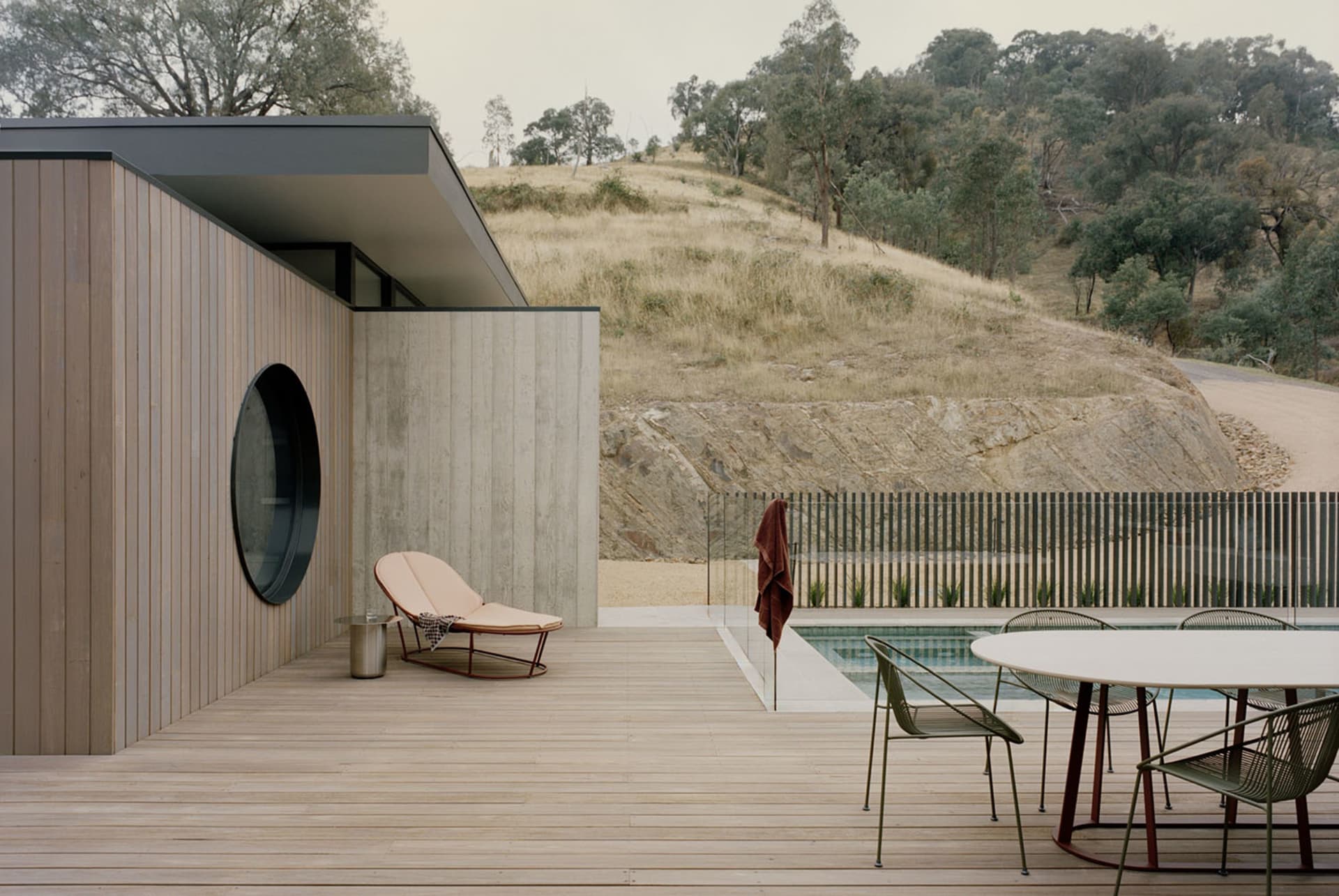 Modern outdoor wooden deck featuring a large circular window, concrete walls, a chaise lounge, and a round dining set beside a swimming pool with a dry hilly landscape in the background.