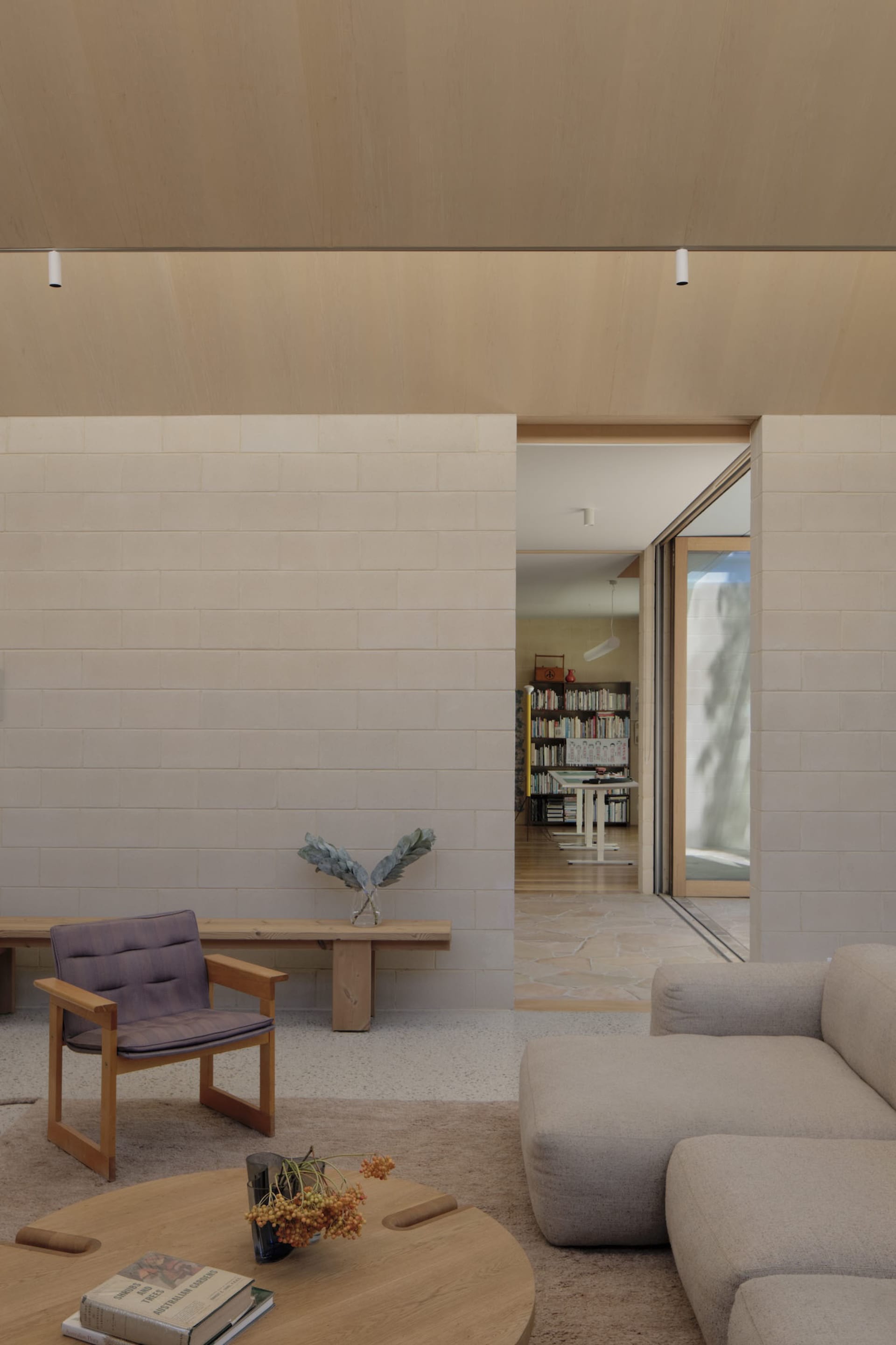Modern minimalist living room featuring white limestone block walls, light wood ceiling, a purple upholstered wooden armchair, and a large beige sectional sofa with a round wooden coffee table in the foreground.