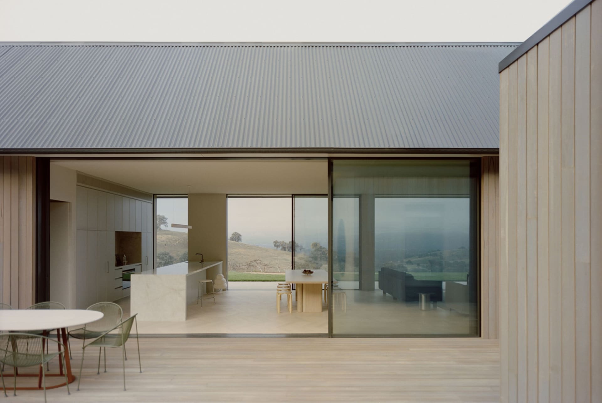 Modern open-plan kitchen and living area with large sliding glass doors opening to rolling countryside hills, featuring a white limestone kitchen island, light oak flooring, and pale green dining chairs.