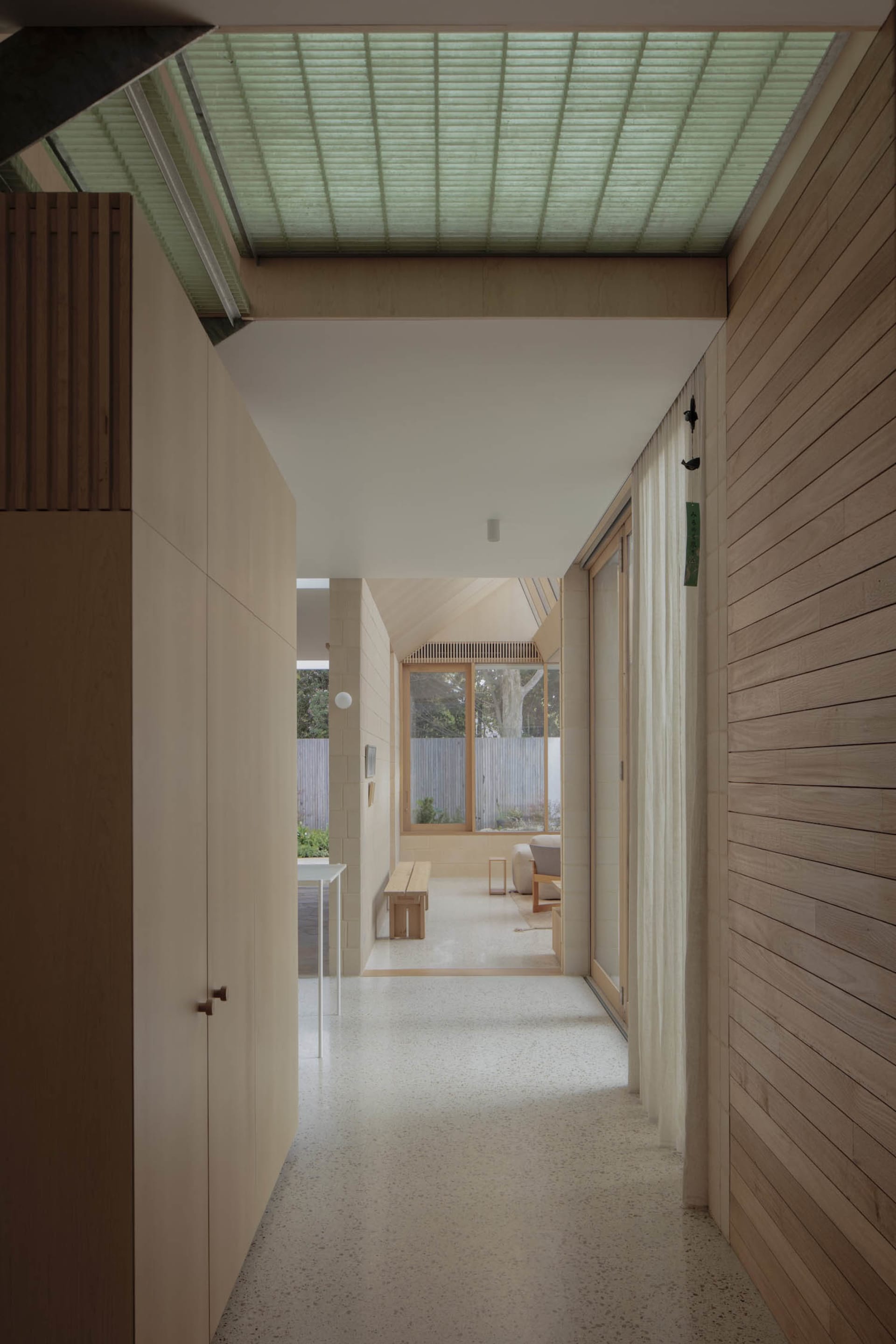 Long modern wooden corridor with light wood paneling, polished terrazzo floor, large clerestory skylight with blinds, and view to a bright garden courtyard.