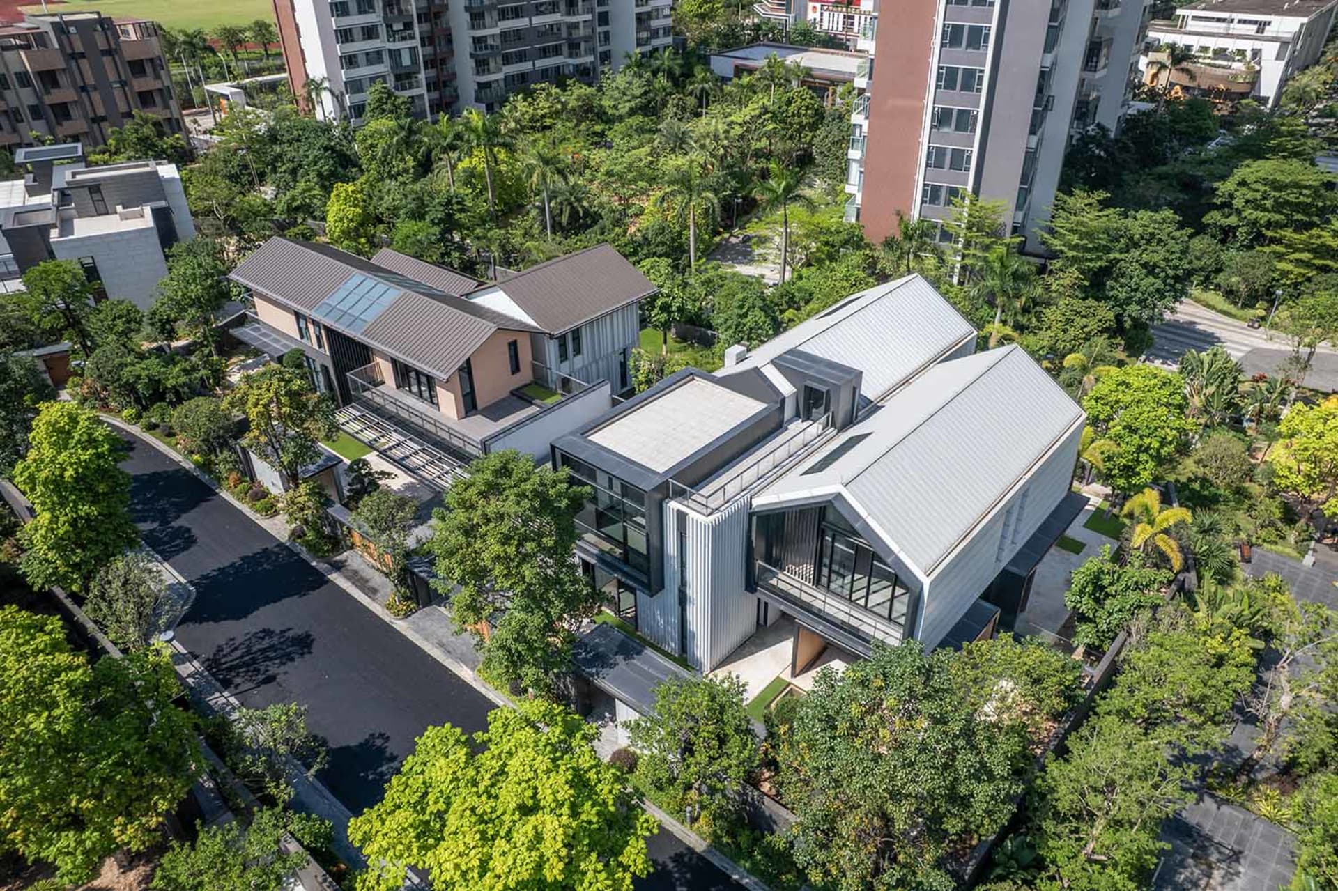 Aerial view of a contemporary luxury villa featuring extensive glass walls, corrugated metal cladding, and flat roofs, nestled among dense tropical trees in a suburban neighborhood.