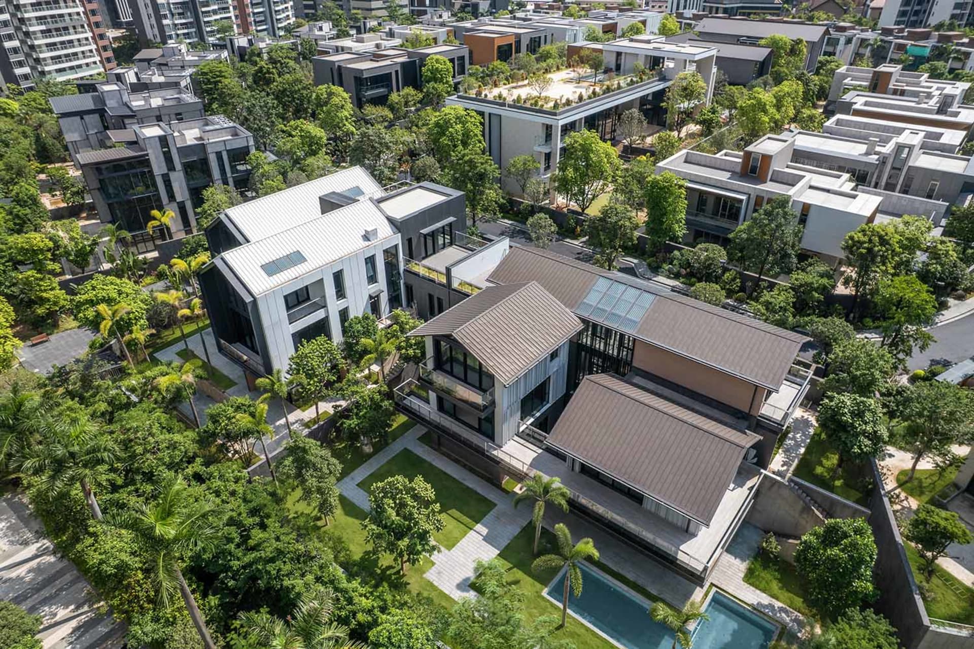 Aerial view of modern luxury villas featuring silver and dark metal cladding, large glass windows, rooftop terraces, private swimming pools, and surrounded by dense tropical greenery in a high-end residential community.