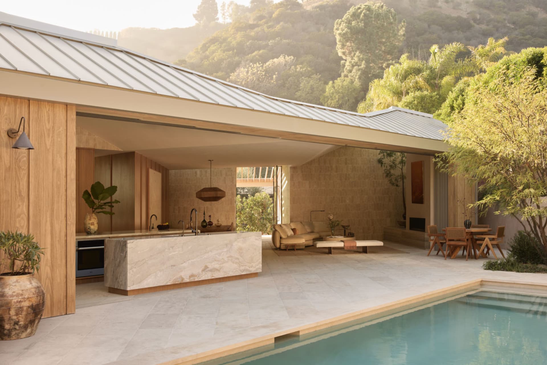 Modern open-plan indoor-outdoor living and kitchen with a large travertine marble island, wooden ceilings, and a swimming pool overlooking a lush hillside.