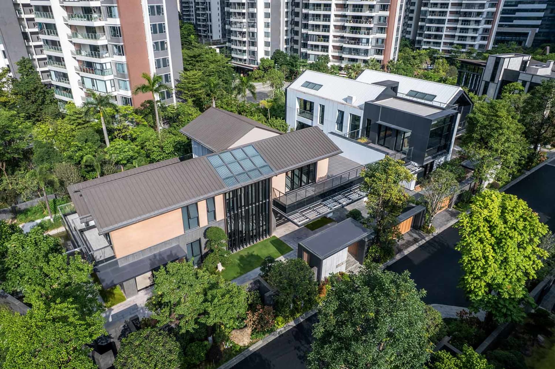 Aerial view of a contemporary two-story villa featuring large glass windows, dark metal roof, warm beige walls, and surrounded by dense green trees and a manicured garden in a suburban setting with apartment buildings in the background.