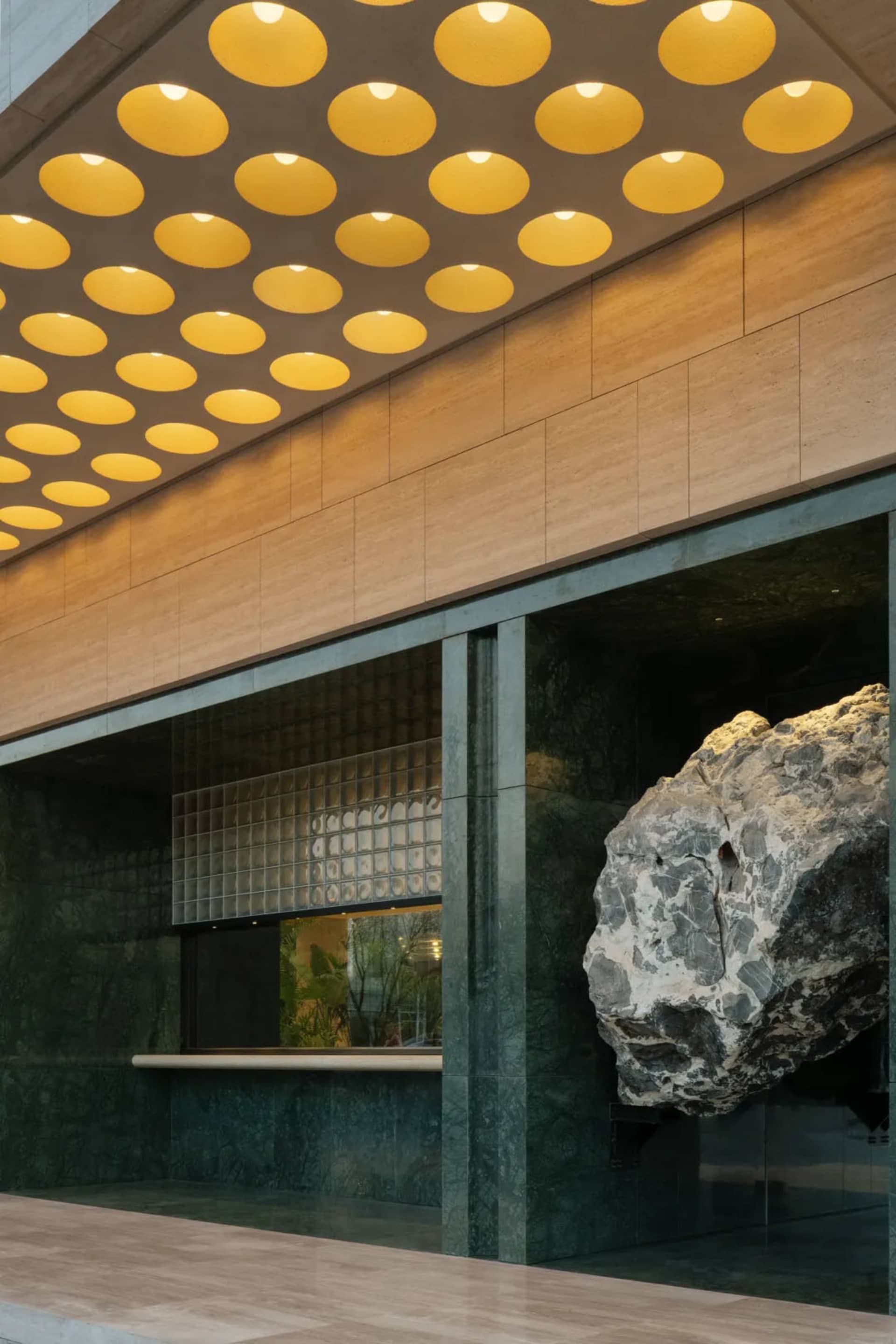 Modern building lobby featuring a large floating granite boulder, green marble walls, travertine ceiling with circular warm lights, and glass block detail.