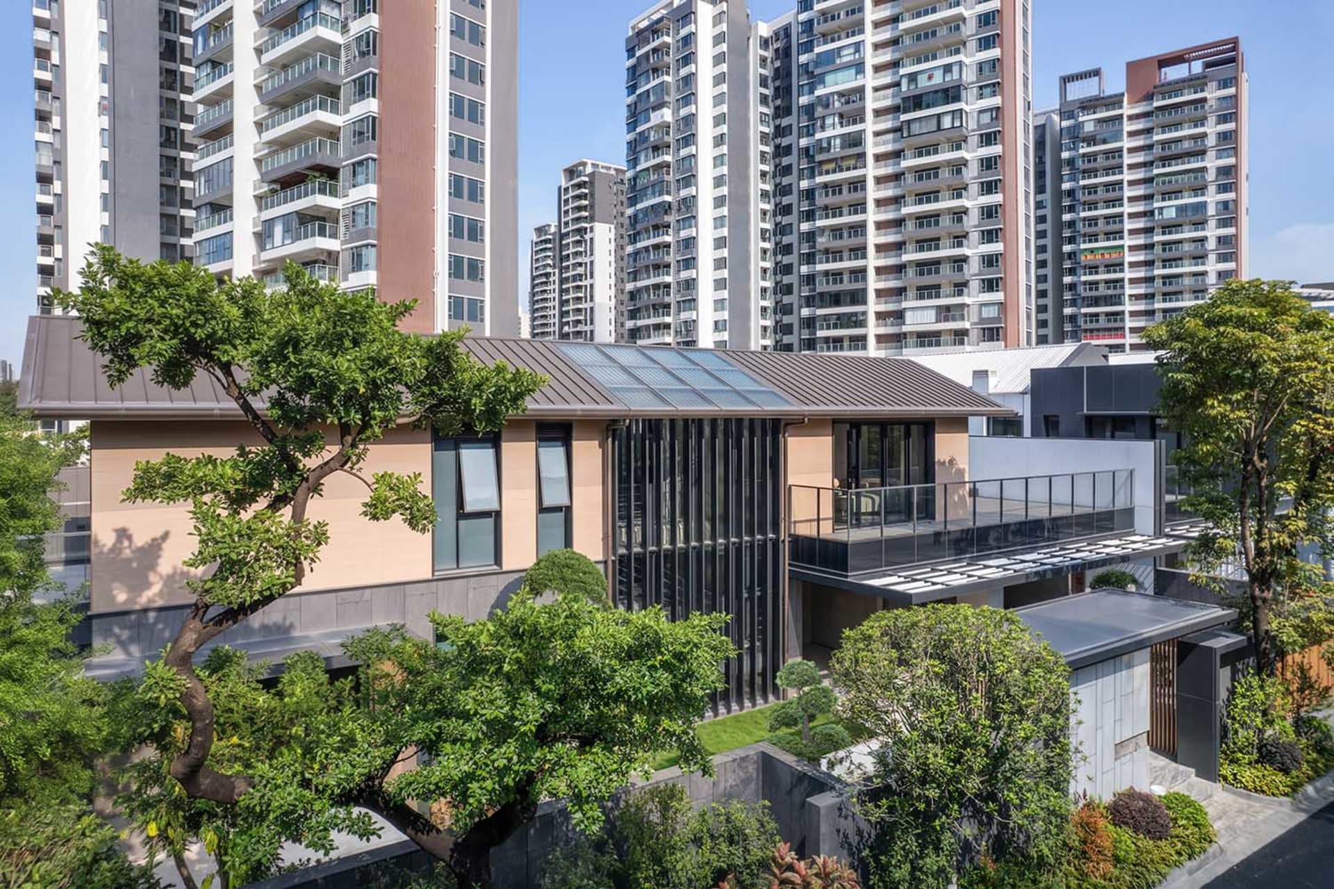 Modern low-rise courtyard house with large glass windows, skylight roof, and lush garden surrounded by tall urban apartment buildings under a clear blue sky.