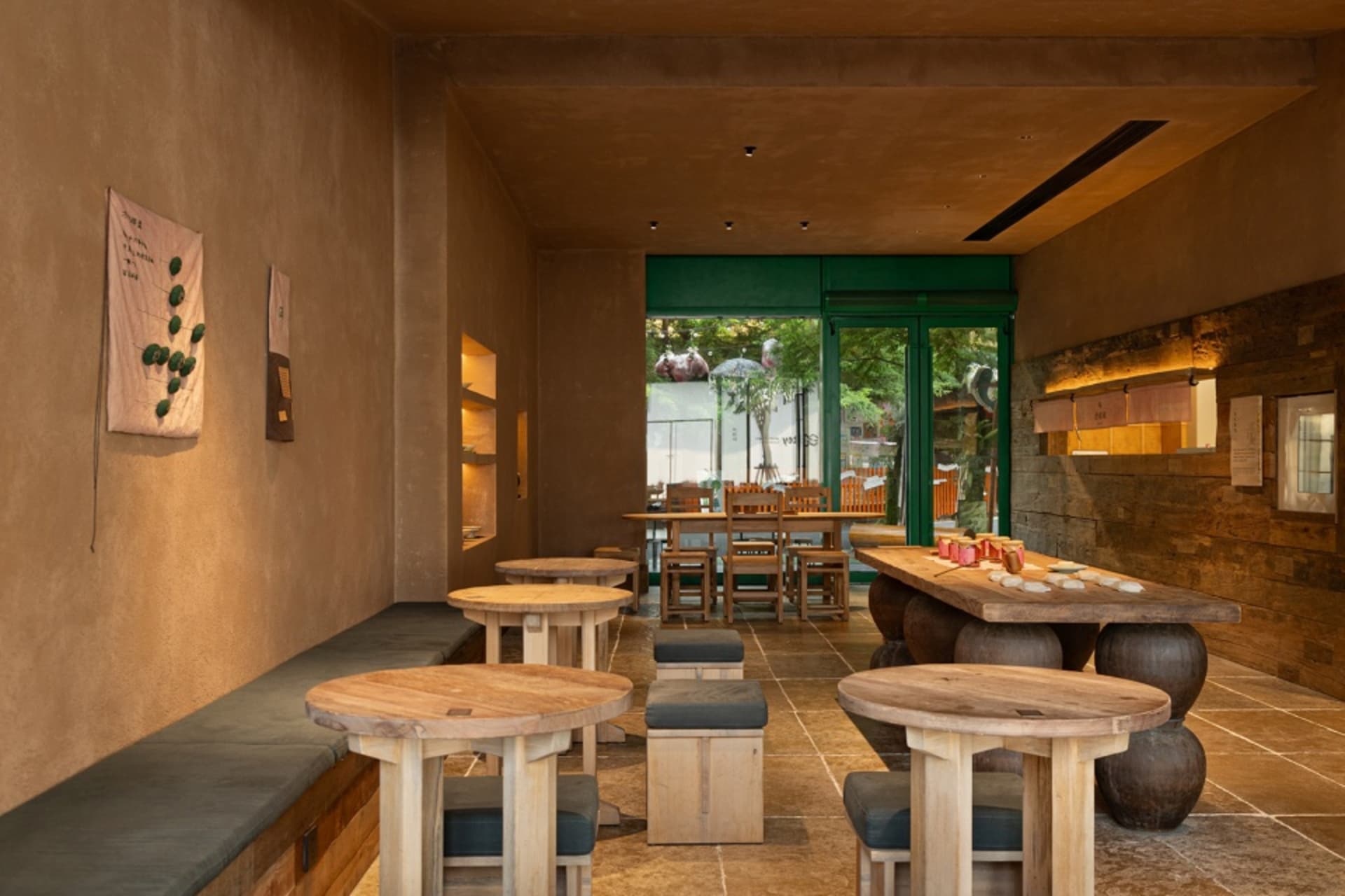Modern wabi-sabi style restaurant interior with round solid wood tables, concrete walls, wooden stools with dark cushions, and large green-framed glass doors opening to a courtyard.