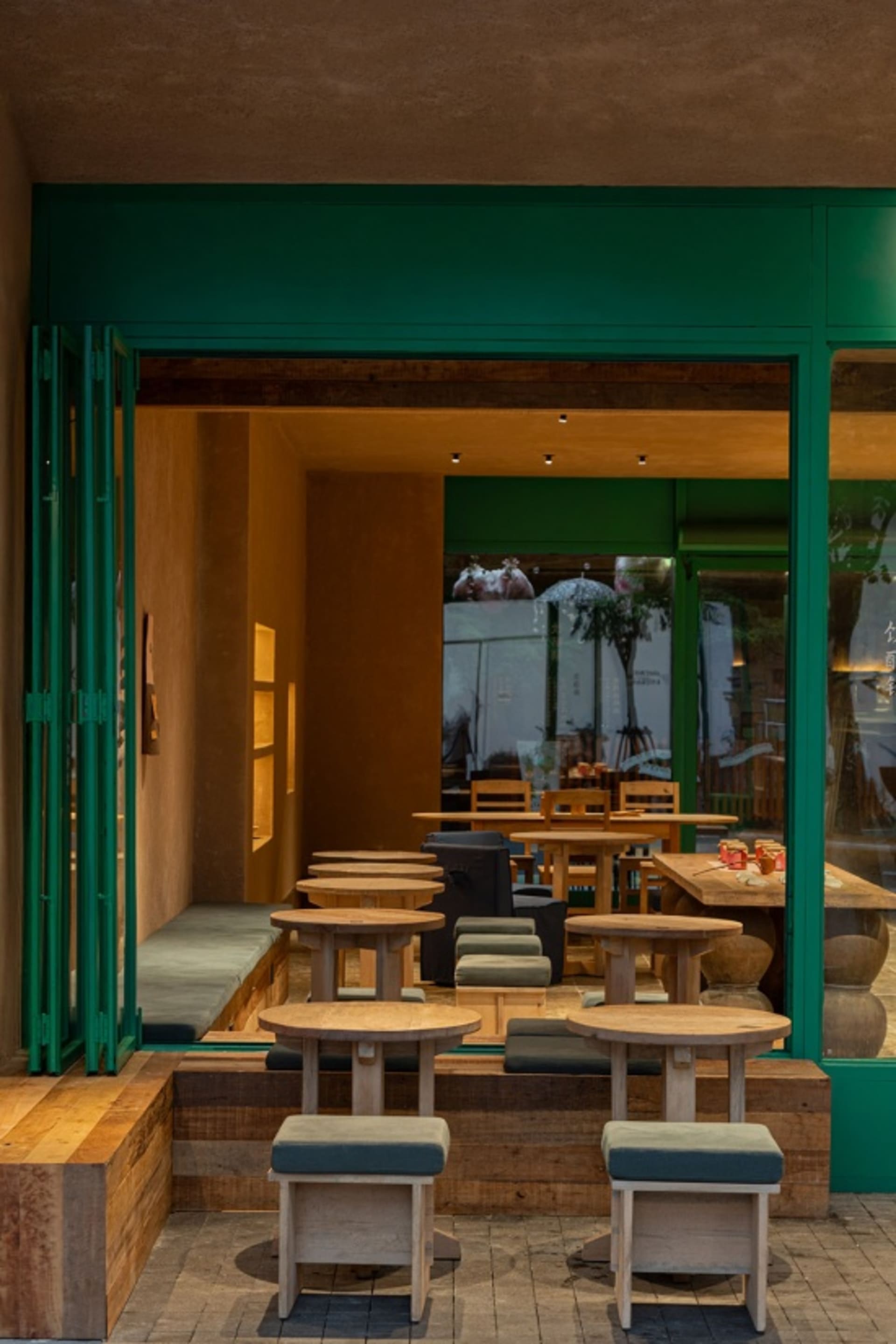 Modern cafe interior featuring round oak stools, built-in wooden banquettes with green cushions, and a bold emerald green entrance frame.