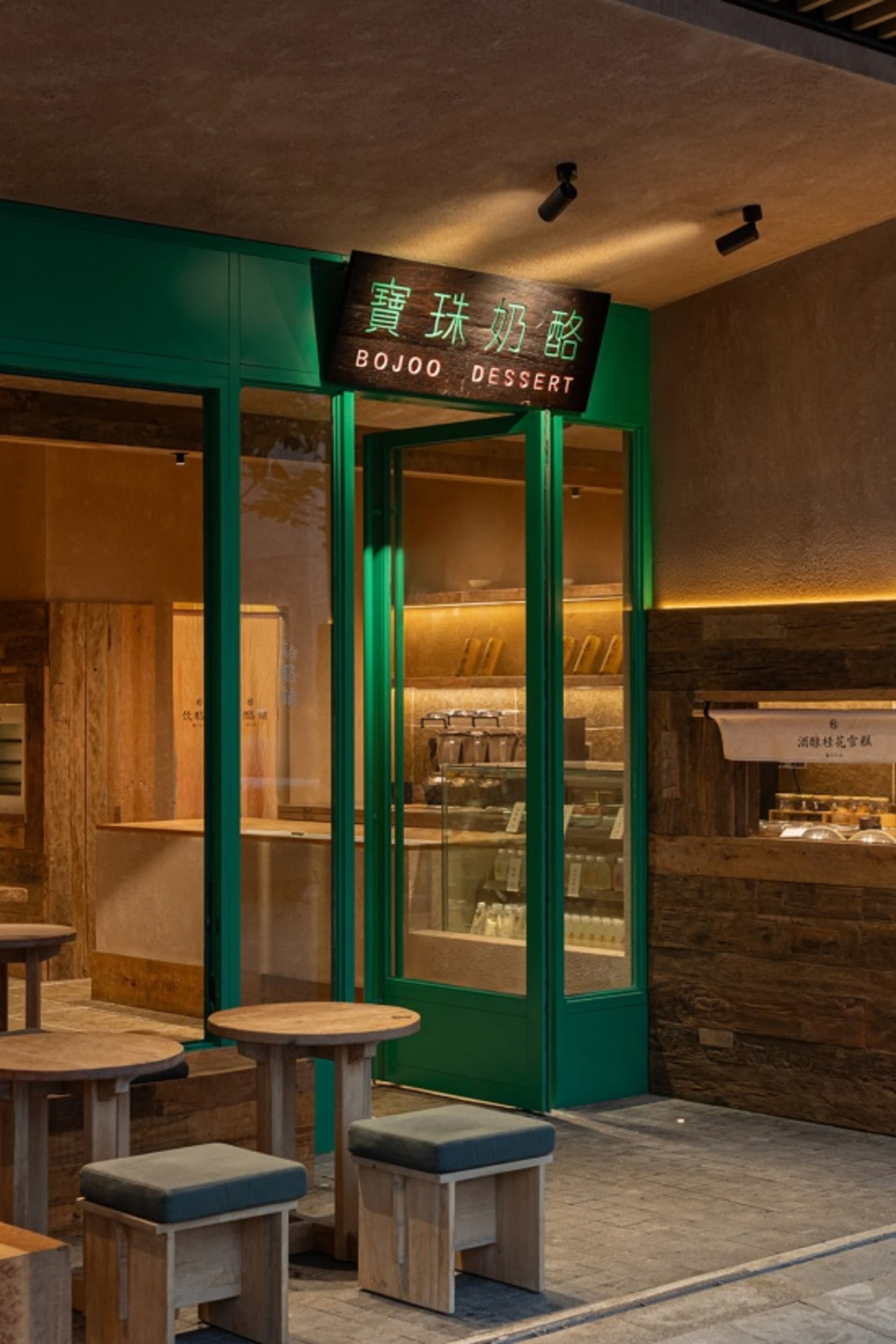 Street view of a modern dessert shop named BOJOO with emerald green door frames, wooden stools and tables in the foreground, and warm interior lighting visible through glass doors.