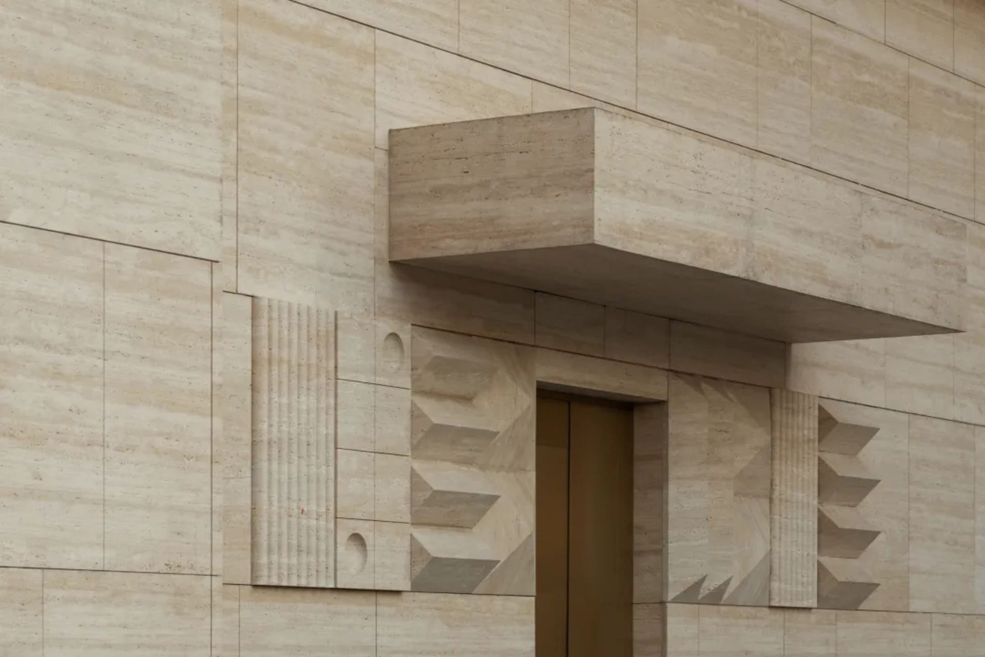 Close-up of a luxurious travertine stone building entrance featuring geometric relief patterns, fluted columns, and a cantilevered overhang above a bronze door.