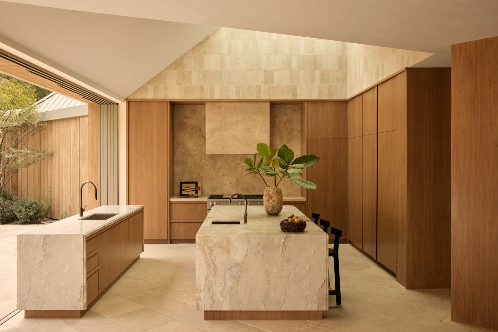 Modern kitchen featuring a large central island made of veined travertine, warm oak cabinetry, black metal chairs, and a tall skylight with textured stone walls.