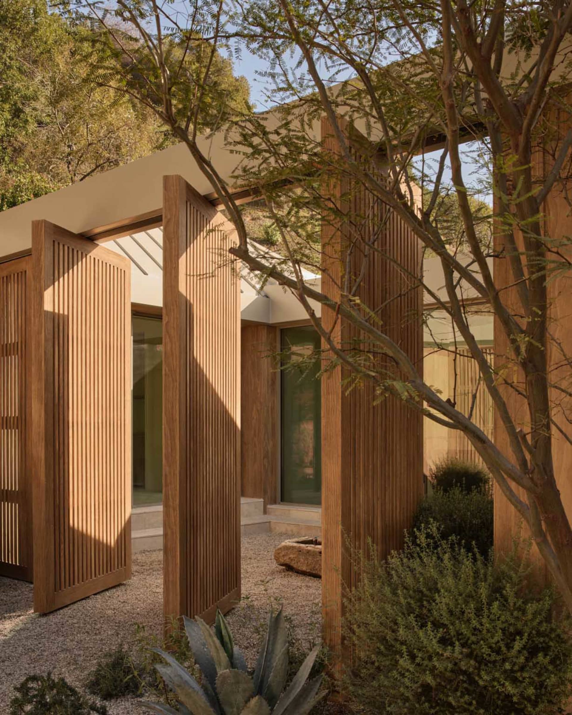 Modern outdoor courtyard featuring vertical teak wood slatted walls and doors, gravel ground, native desert plants, and a mountain backdrop under a clear blue sky.