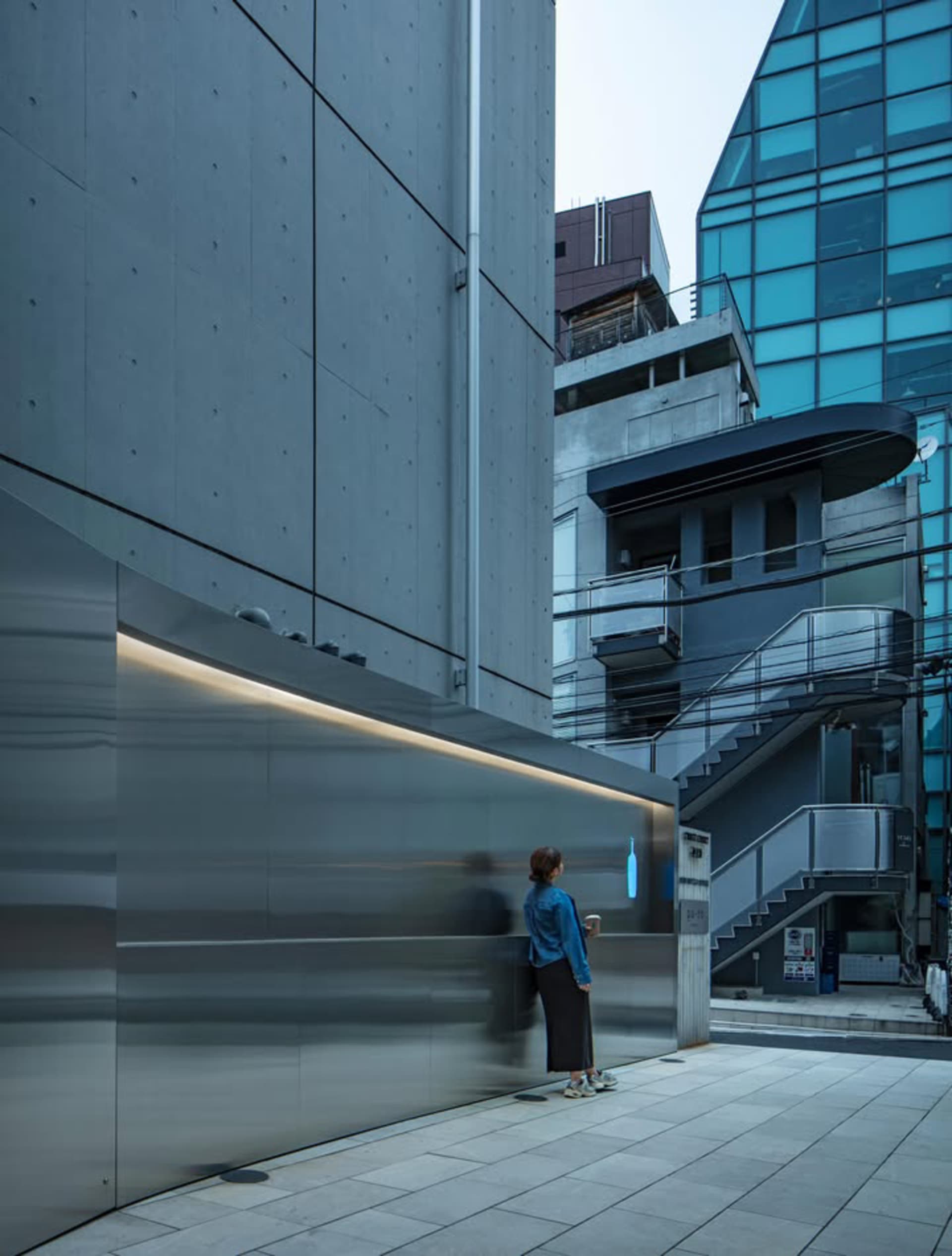 Woman standing next to a curved stainless steel wall with integrated lighting in a modern urban alley surrounded by brutalist concrete buildings.