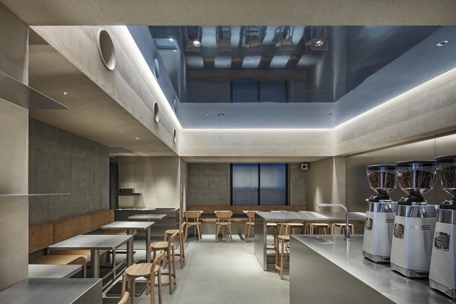 Modern minimalist café interior featuring raw concrete walls, stainless steel counters, large coffee grinders, and light oak wooden stools and tables under dramatic linear ceiling lighting.