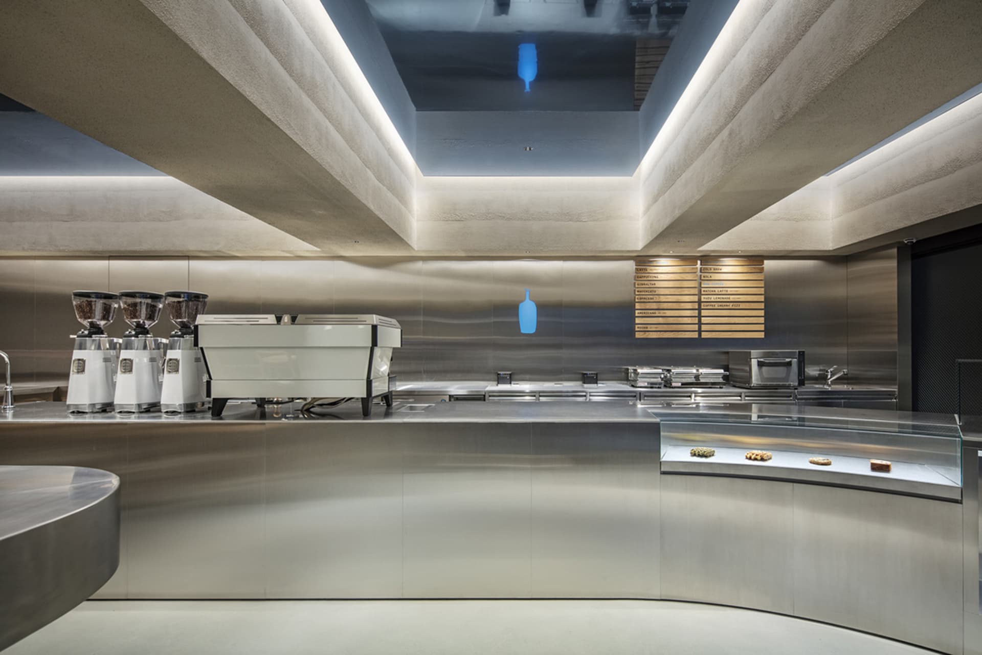 Modern minimalist coffee shop service counter made of curved stainless steel with concrete ceiling and integrated LED lighting