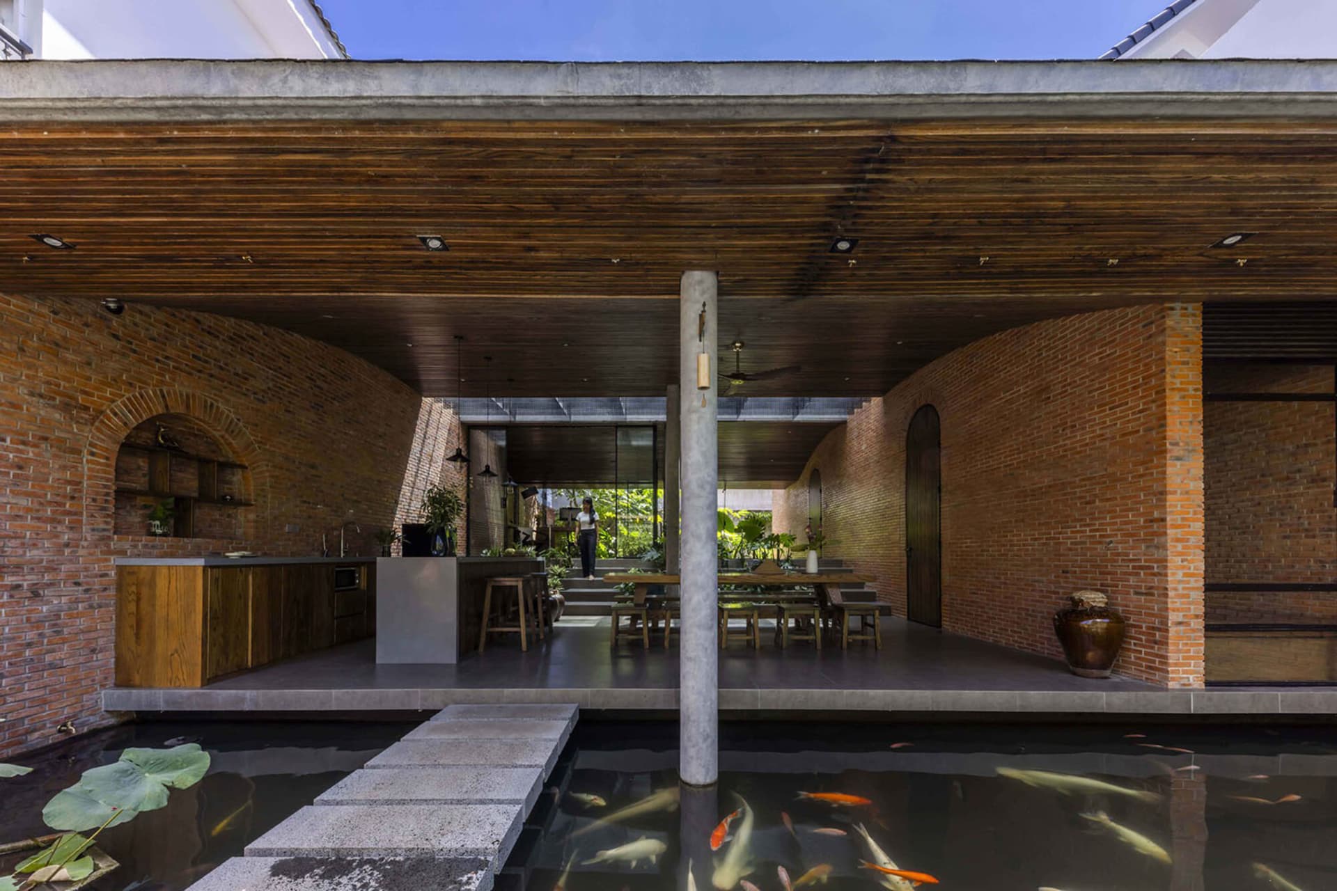 Modern tropical courtyard house featuring curved exposed brick walls, wooden ceiling, open kitchen, dining area, and a foreground koi pond with stepping stones.