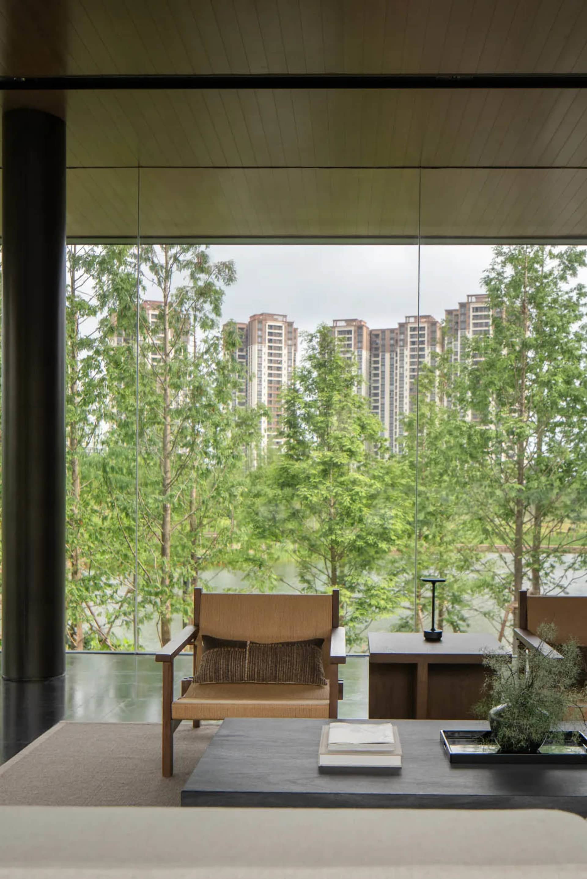 Modern minimalist living room featuring wooden armchairs, dark stone coffee table, and expansive glass windows revealing a green lakeside view with city buildings in the background.
