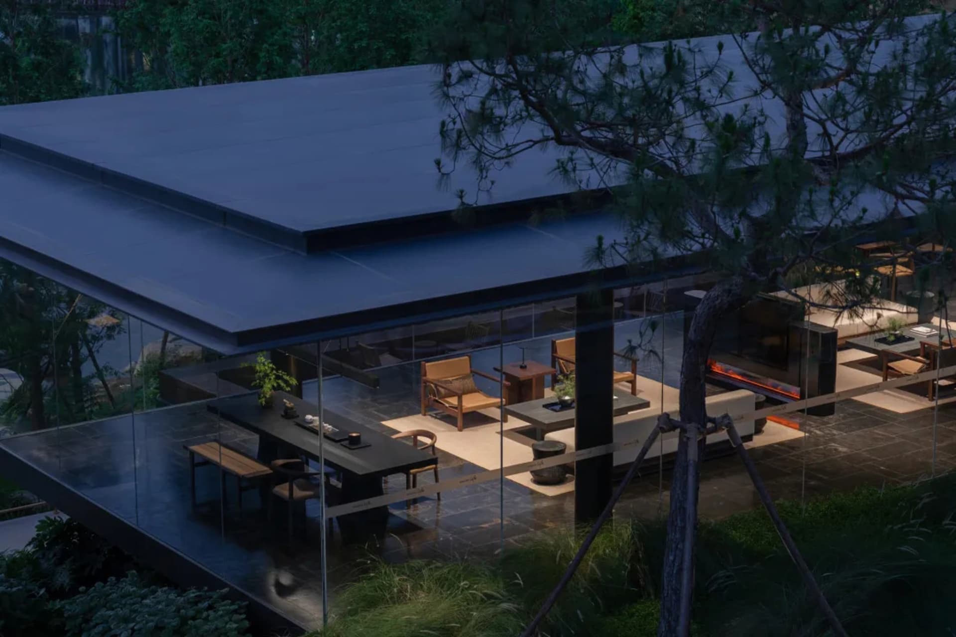 Night view of a modern glass villa with an open terrace featuring wooden furniture, a long dining table, and a fireplace, overlooking lush greenery with a prominent pine tree in the foreground.