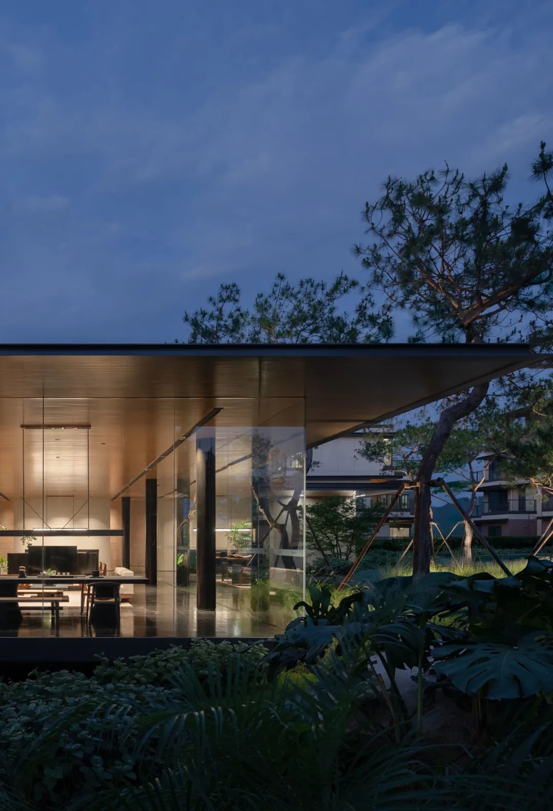 Modern glass villa with warm interior lighting at dusk, surrounded by tropical plants and pine trees in a landscaped garden.