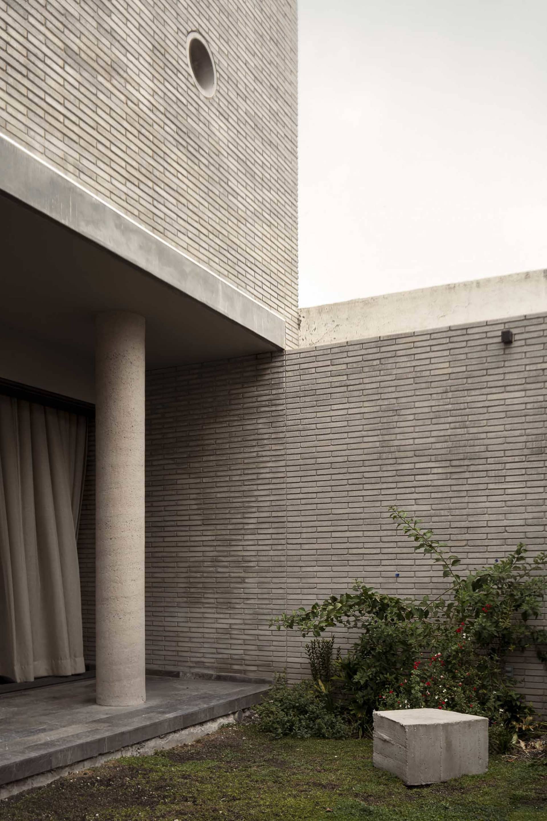 Exterior view of a modern brutalist courtyard featuring an exposed concrete column, grey brick walls, a circular window, and lush greenery with a concrete block stool.