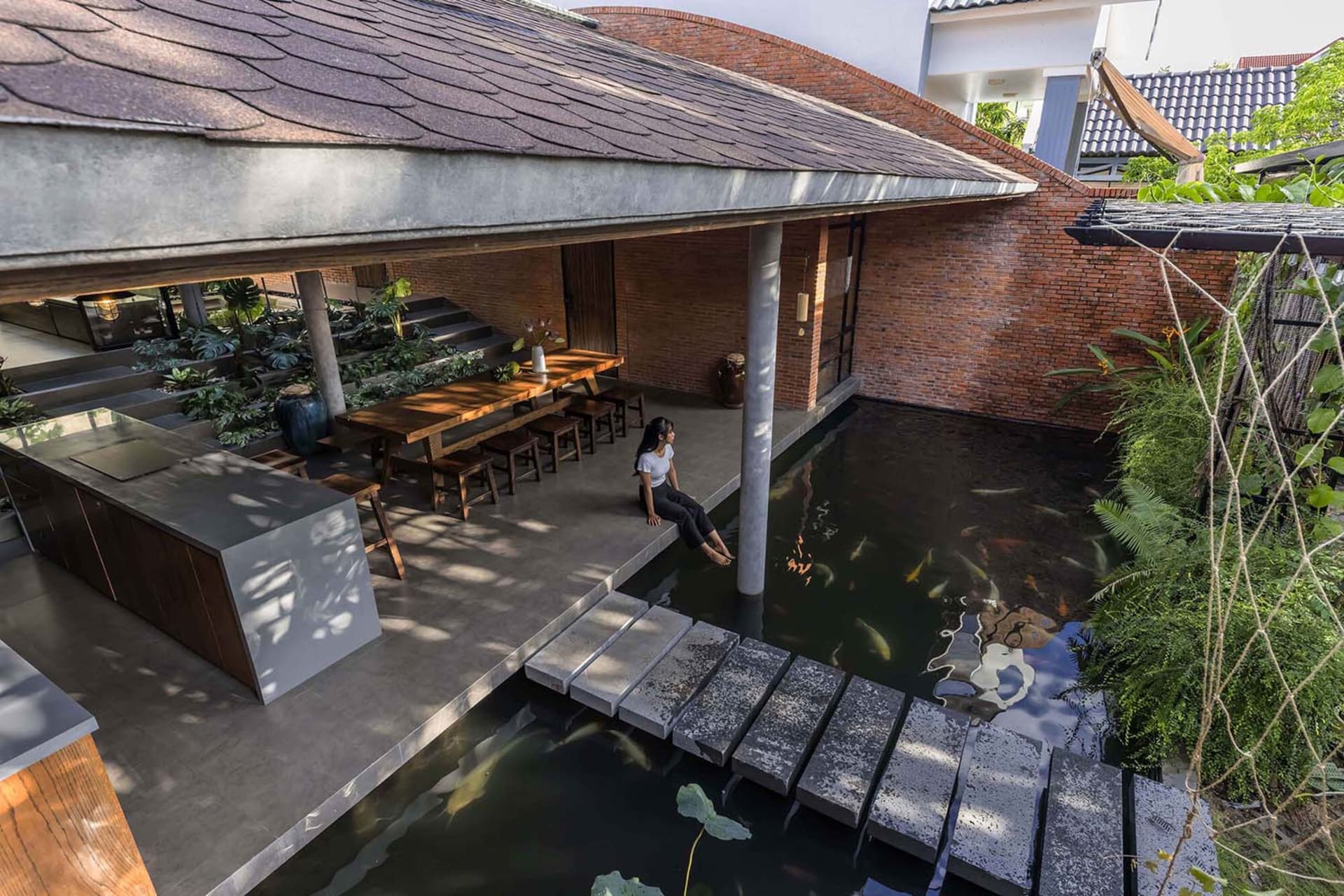 High-angle view of a modern tropical courtyard house featuring a large koi pond, exposed red brick walls, concrete walkway, wooden dining table, and lush greenery.