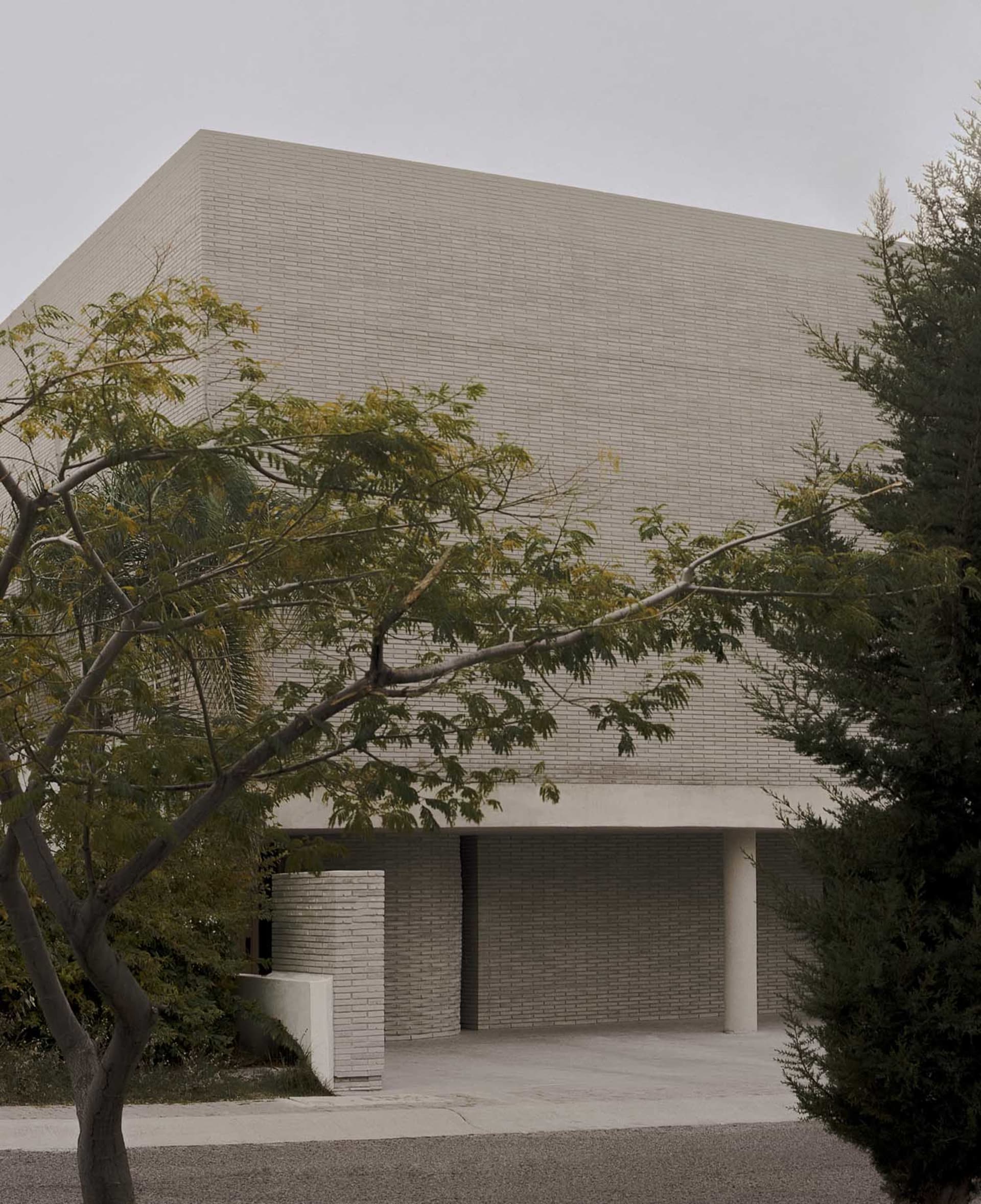 Exterior view of a brutalist concrete building with a strong geometric form, overhanging canopy, and surrounding trees under an overcast sky.