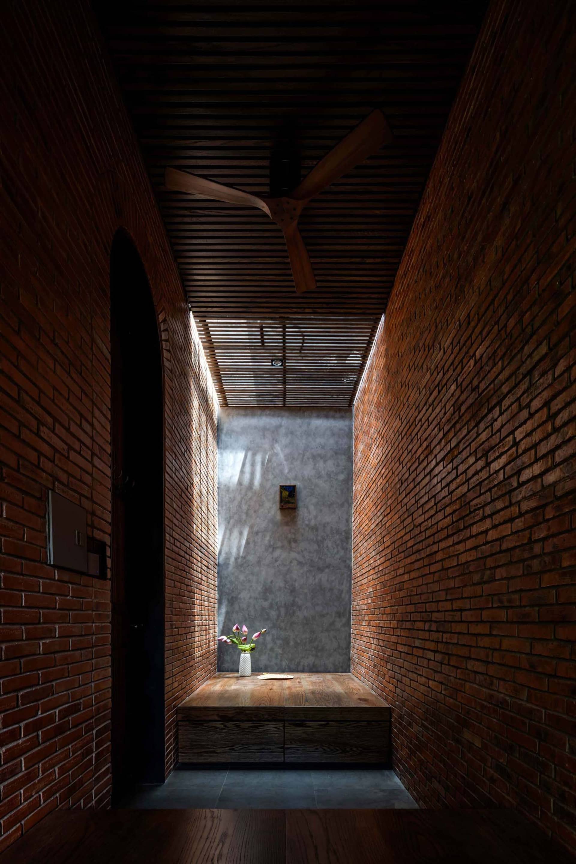 Narrow architectural hallway with red brick walls, wooden slat ceiling, built-in wooden bench, and a concrete end wall with natural light from a skylight.