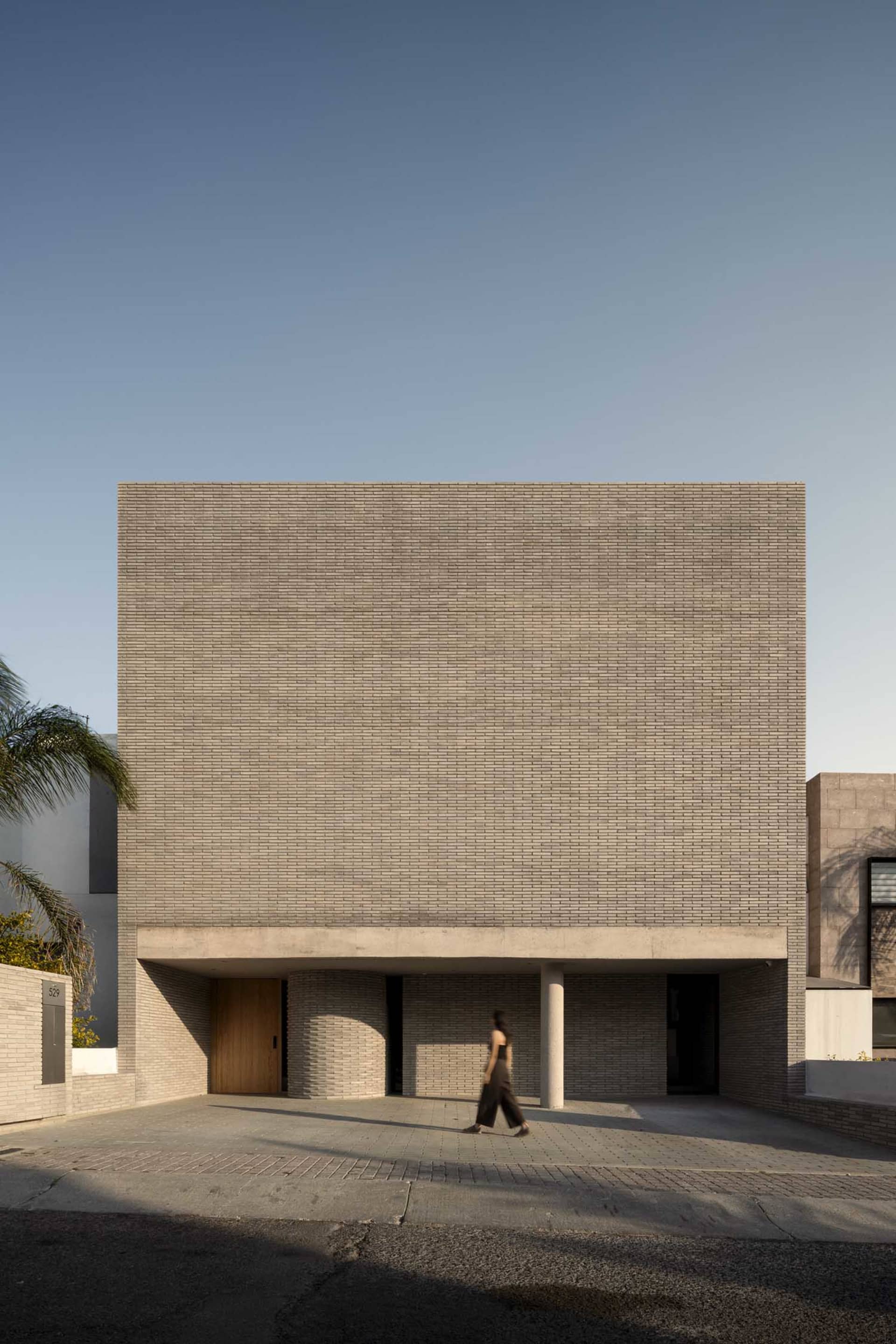 Front view of a modern brutalist brick building with a wooden door, cylindrical columns, and a woman walking across the paved courtyard under clear blue sky.