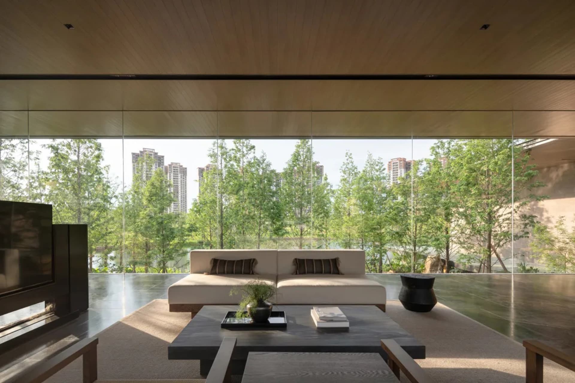 Modern minimalist living room featuring a low-profile cream sofa, dark stone coffee table, wood-paneled ceiling, and expansive floor-to-ceiling windows overlooking lush green trees and city buildings.