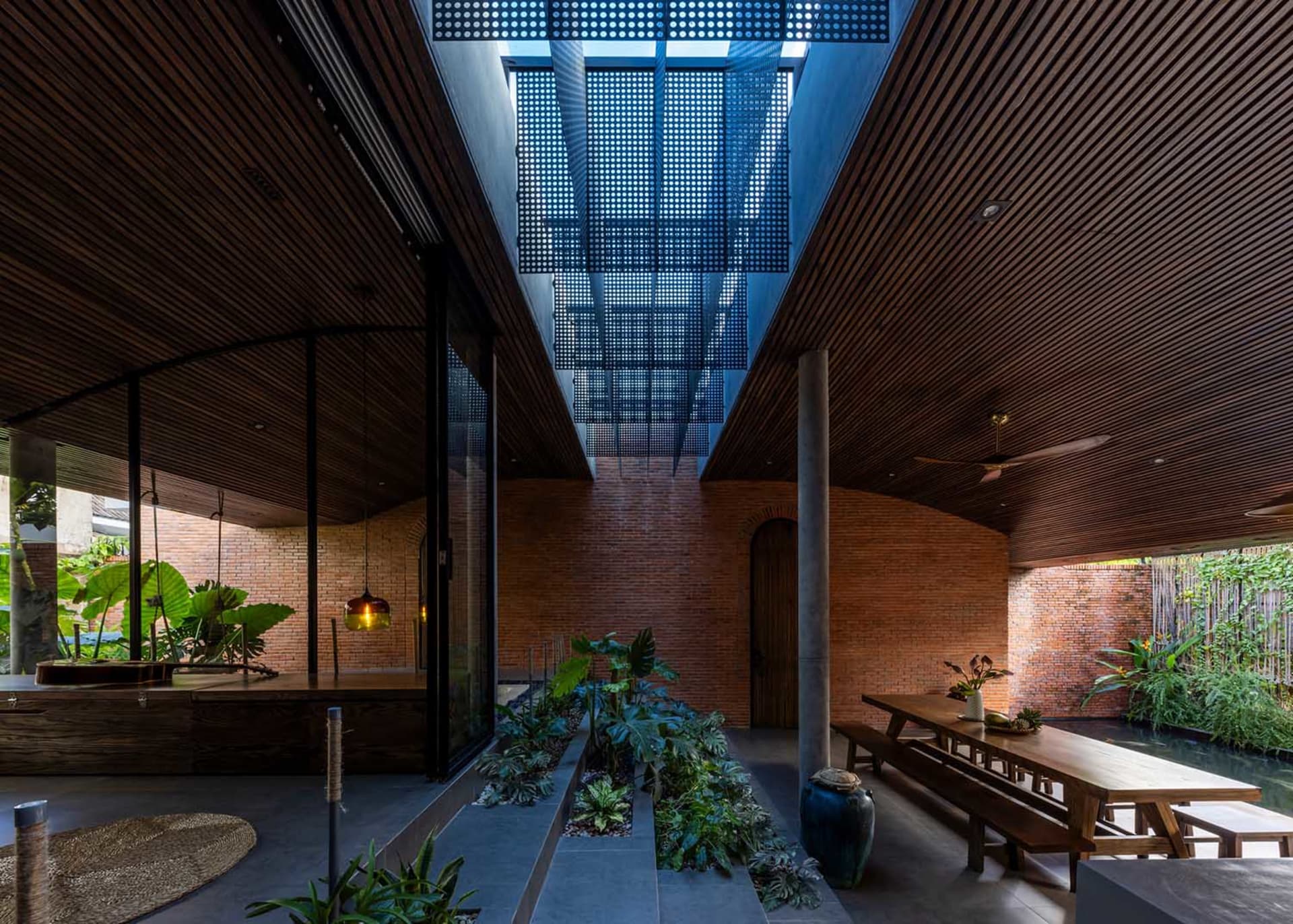 Modern tropical indoor courtyard with exposed red brick walls, dark wooden slat ceiling, central greenery, long wooden dining table, and a large skylight with perforated panels.
