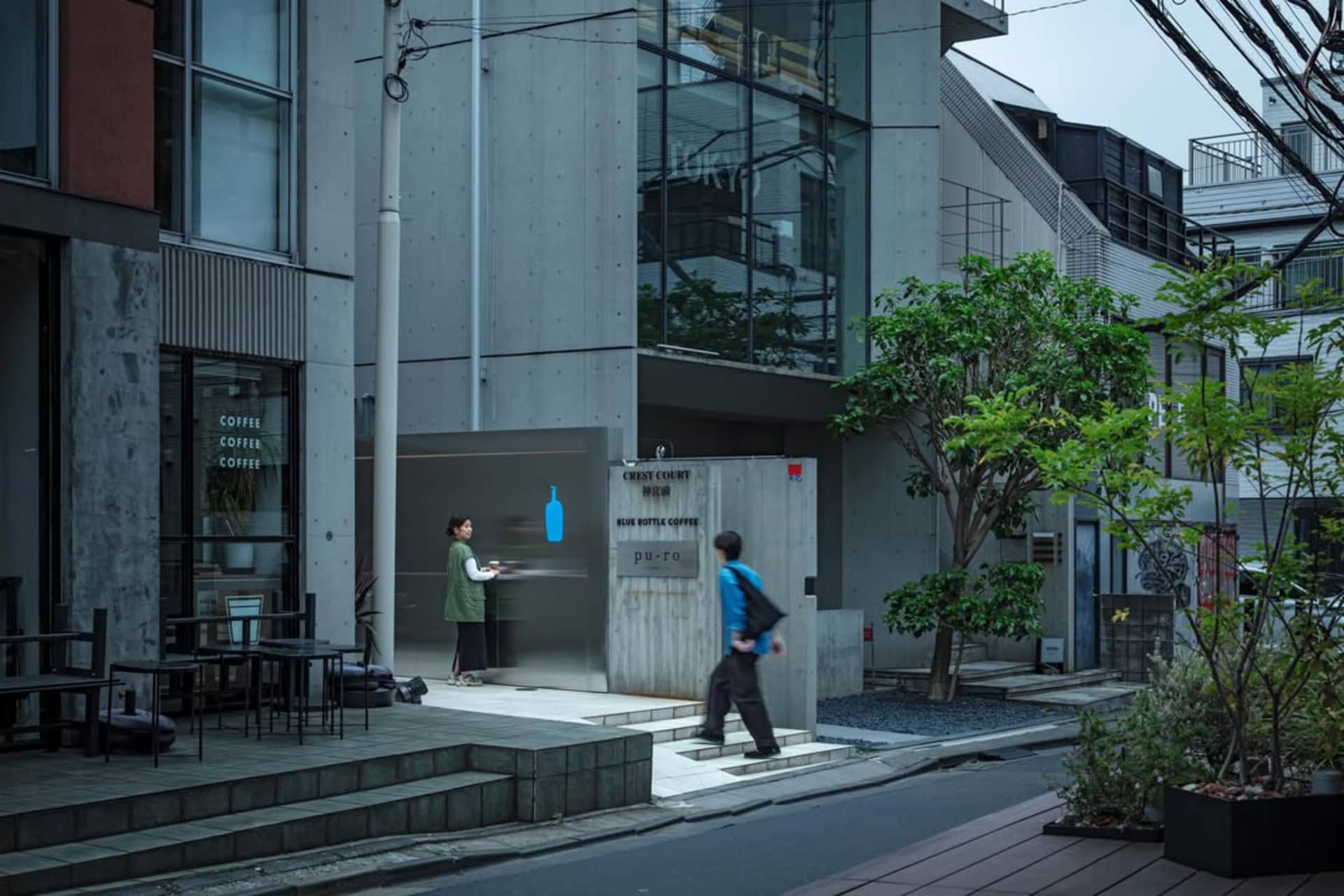 Modern minimalist concrete cafe facade with a woman in a green vest standing at the counter and a pedestrian walking by on a quiet Taipei street.