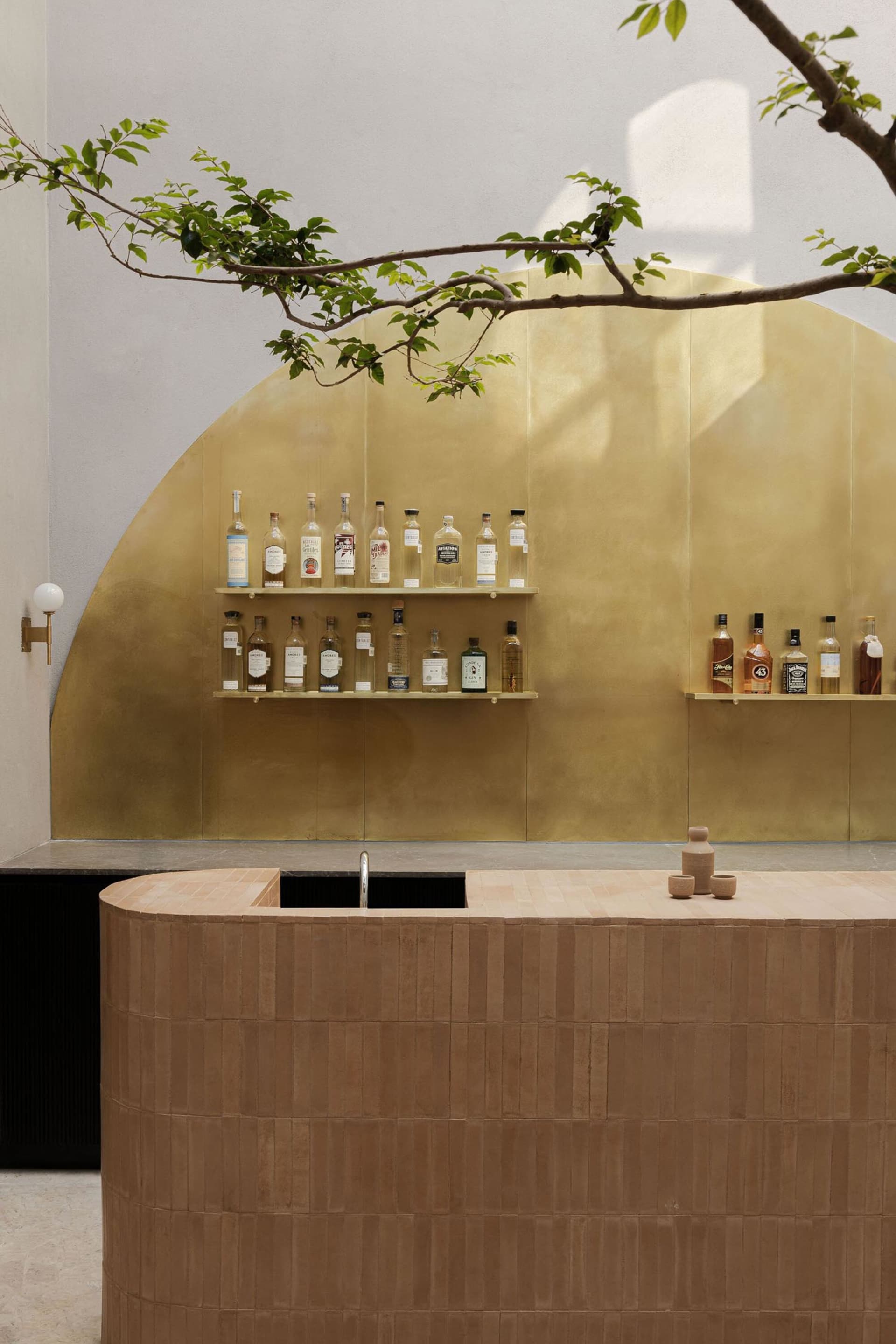 Modern home bar with a curved terracotta-tiled counter, integrated sink, floating shelves displaying liquor bottles against a large brass arched wall panel, with a tree branch overhead.