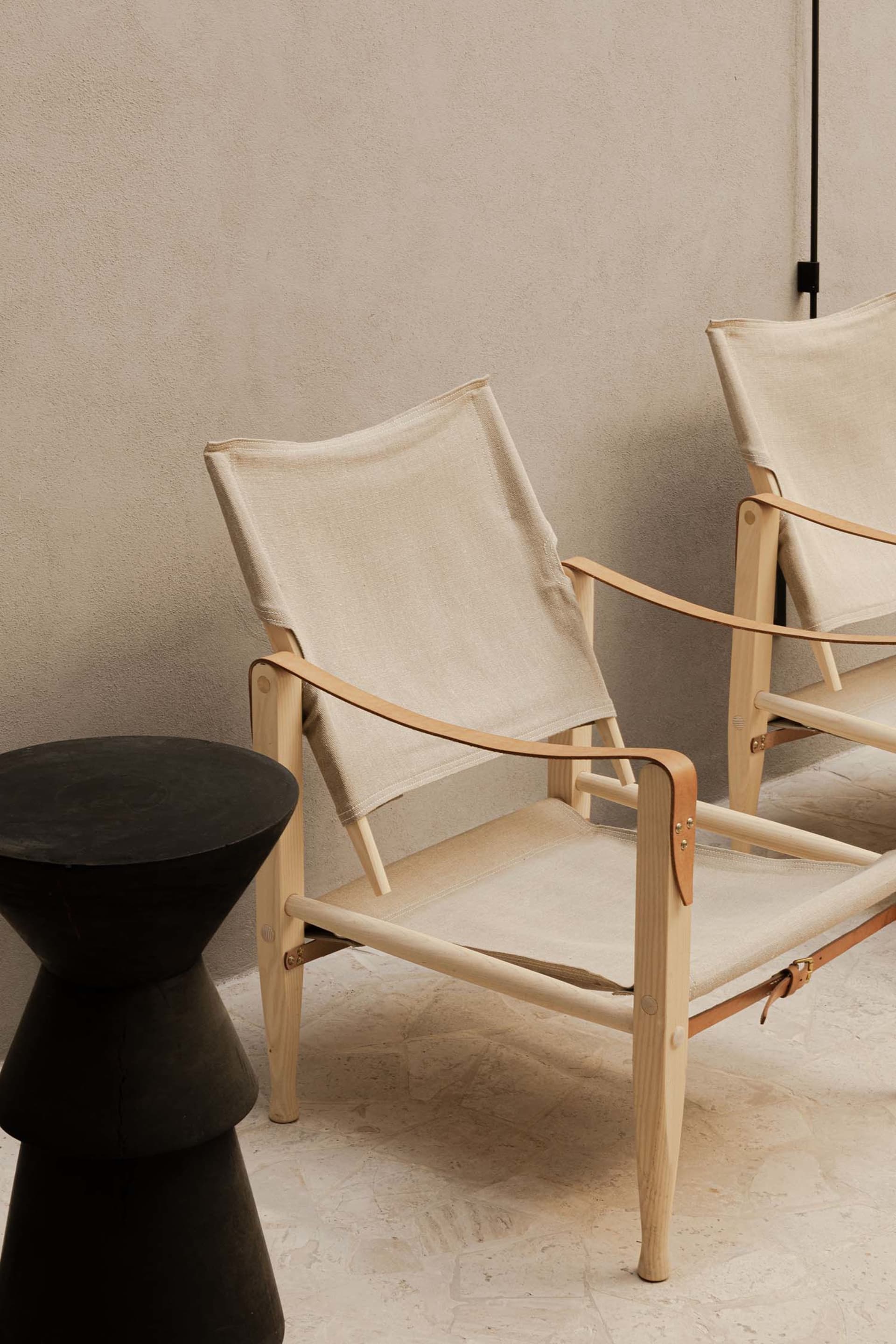 Two minimalist lounge chairs with light ash wood frames and off-white canvas seats and backs, paired with a black sculptural side table against a textured neutral wall.