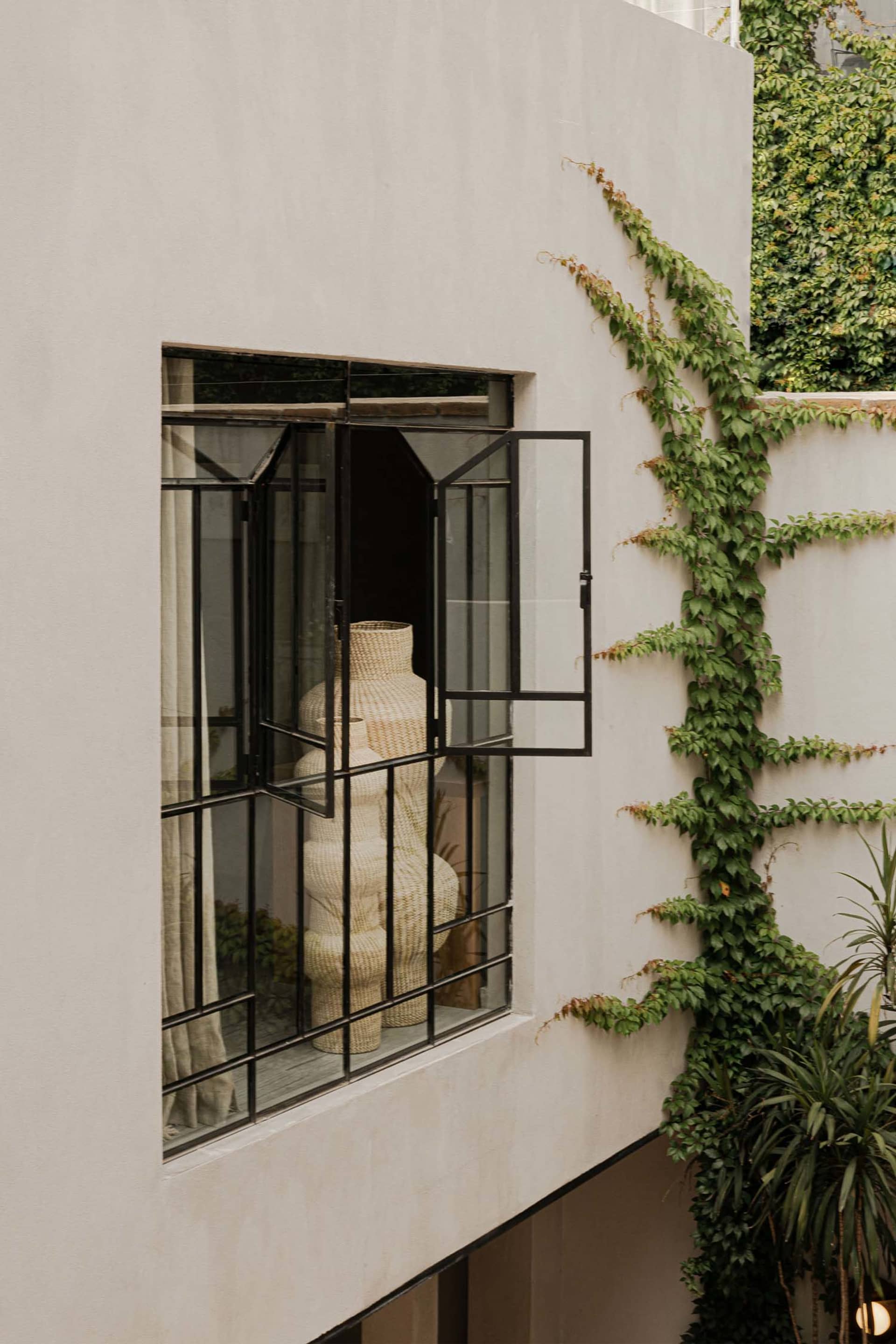 Modern minimalist exterior wall featuring a large black steel pivot window open to reveal tall woven rattan vases inside, with green ivy climbing the adjacent white concrete wall and potted plants below.