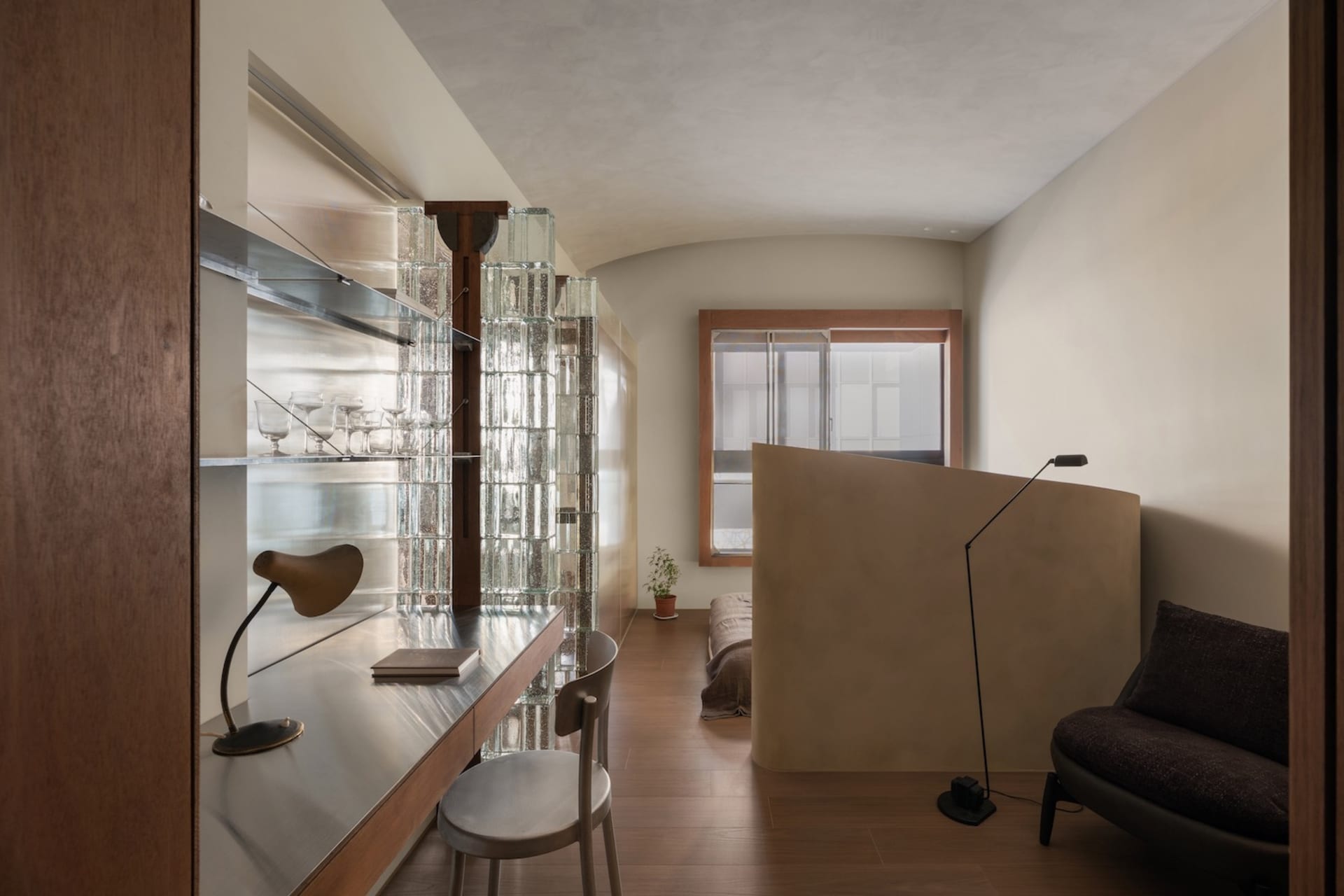 Modern compact studio apartment featuring a built-in stainless steel desk, wooden chair, glass block partition wall, curved rattan room divider, and a daybed in the background.