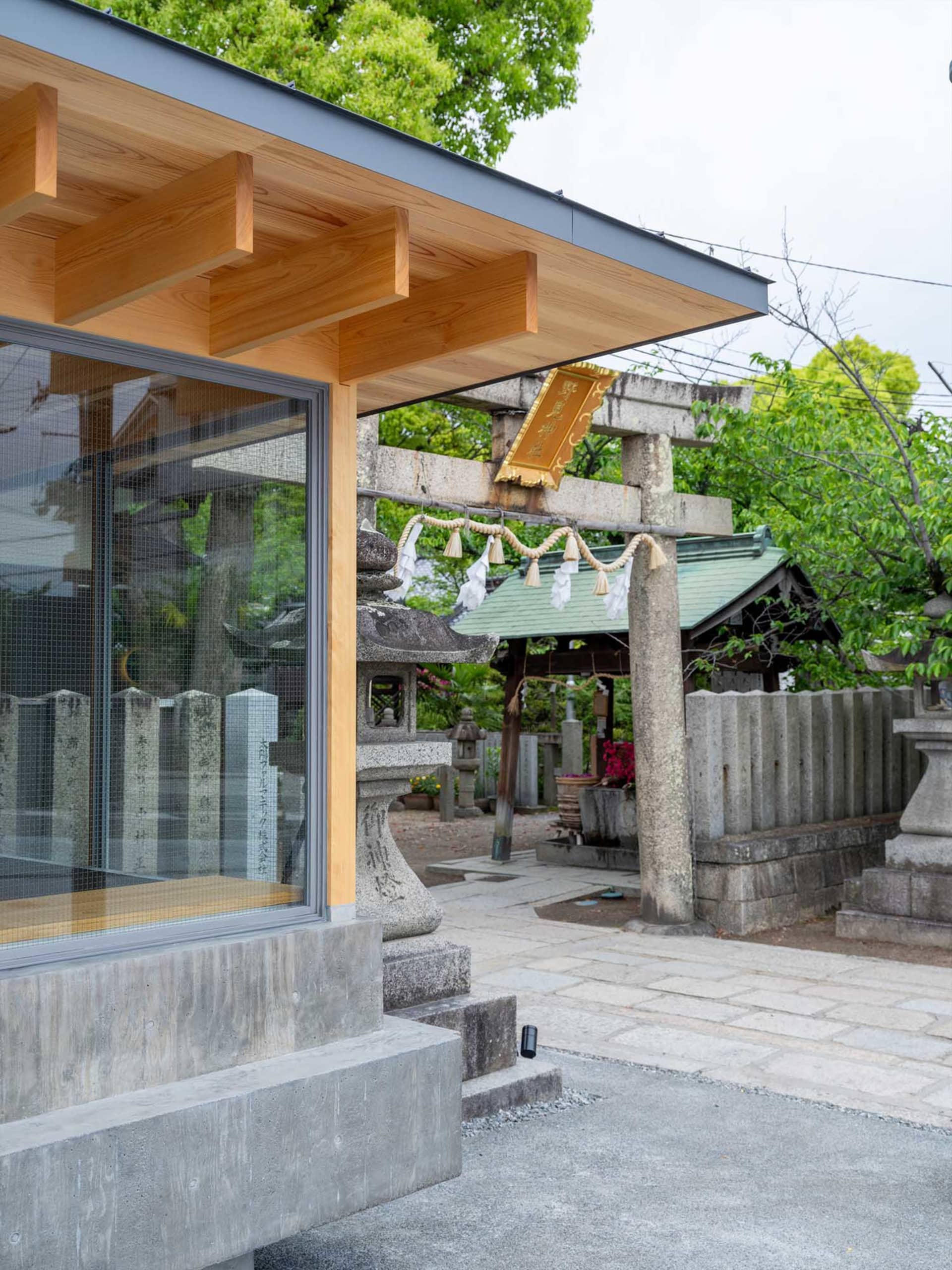 Modern Japanese shrine entrance featuring dramatic cantilevered wooden eaves, concrete steps, stone torii gate, and traditional stone lanterns under lush green trees.
