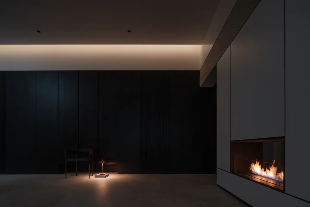 Dark minimalist living room in the evening layered with ceiling cove lighting, a warm table lamp, and a glowing fireplace.