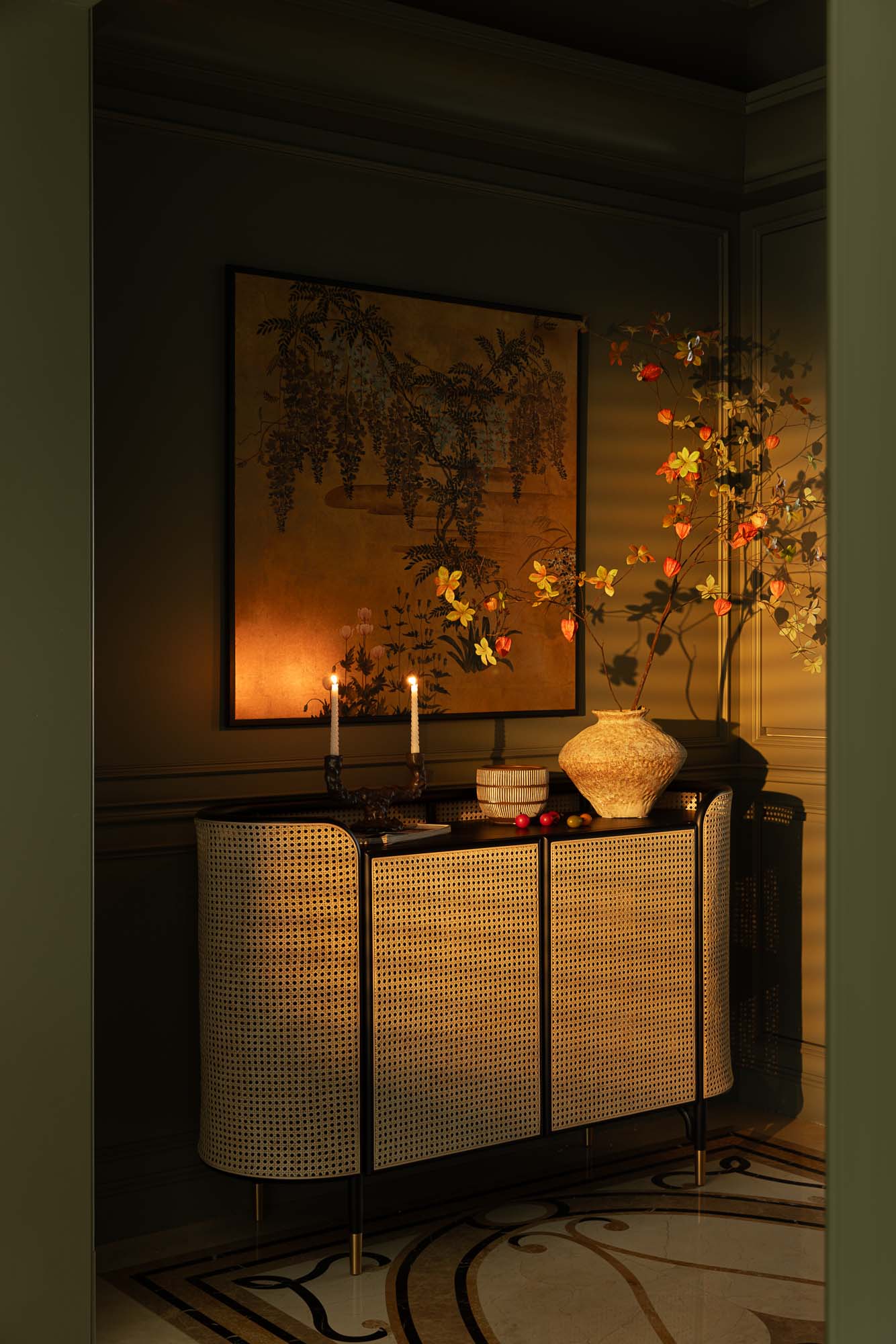 Two lit taper candles and autumnal branches glow warmly on a cane cabinet in a dimly lit room.