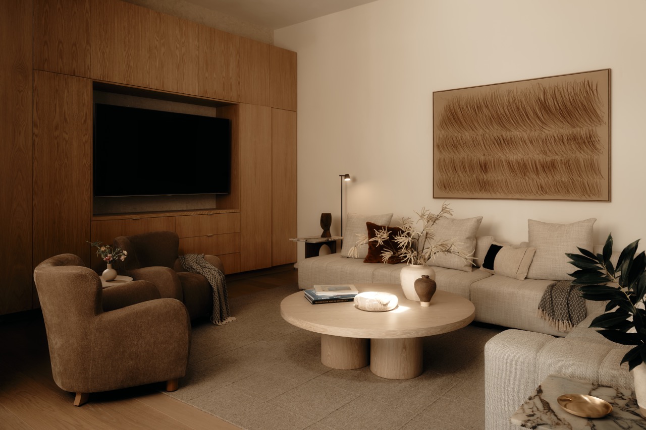 Small warm-toned living room in the evening with a plush armchair, beige sofa, wood cabinetry, and floor lamp glow.