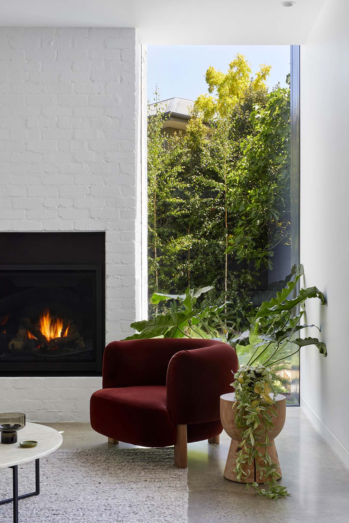 Bordeaux velvet armchair in a cozy reading corner beside a lit fireplace and tall window with garden view.