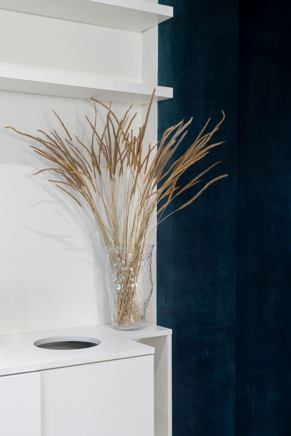 Tall dried pampas grass stems in a glass vase styled on a white shelf against a dark blue curtain.