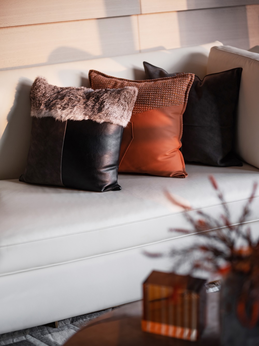 Close-up of textured cushions in terracotta, faux fur, and dark leather on a beige sofa with dried branches nearby.